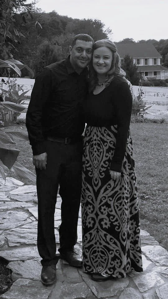 A man and woman standing close together outdoors, both smiling at the camera. The man is wearing a dark shirt and pants, and the woman is wearing a long sleeve sundress. They are on a stone pathway with a house and trees in the background.