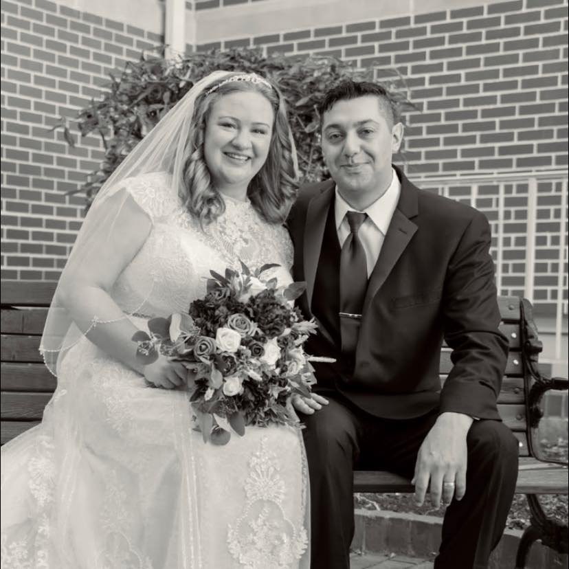 A black and white photo of a bride and groom sitting on a park bench. The bride is dressed in a wedding gown with lace details, wearing a veil and holding a bouquet of flowers. The groom is in a dark suit, white shirt, and tie. They are smiling and posed close together, with a brick wall and some foliage in the background.