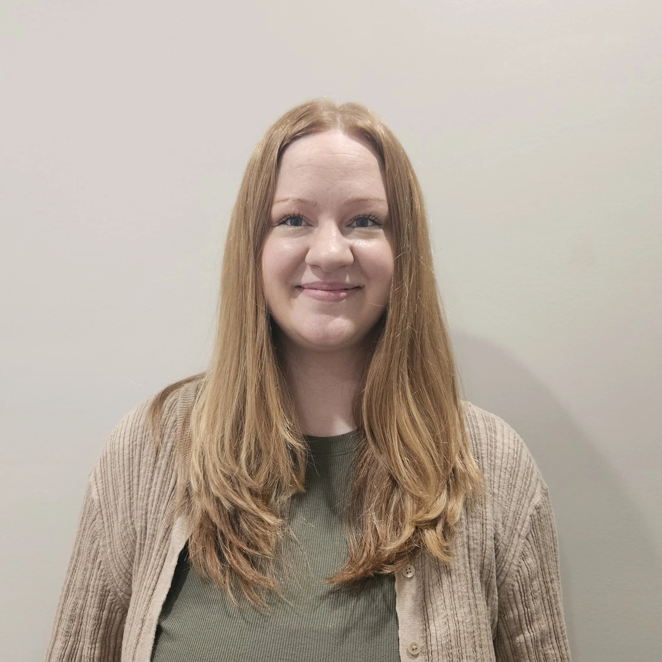 Close-up of a woman with red hair smiling, wearing a beige cardigan over a green top, against a plain off-white wall.