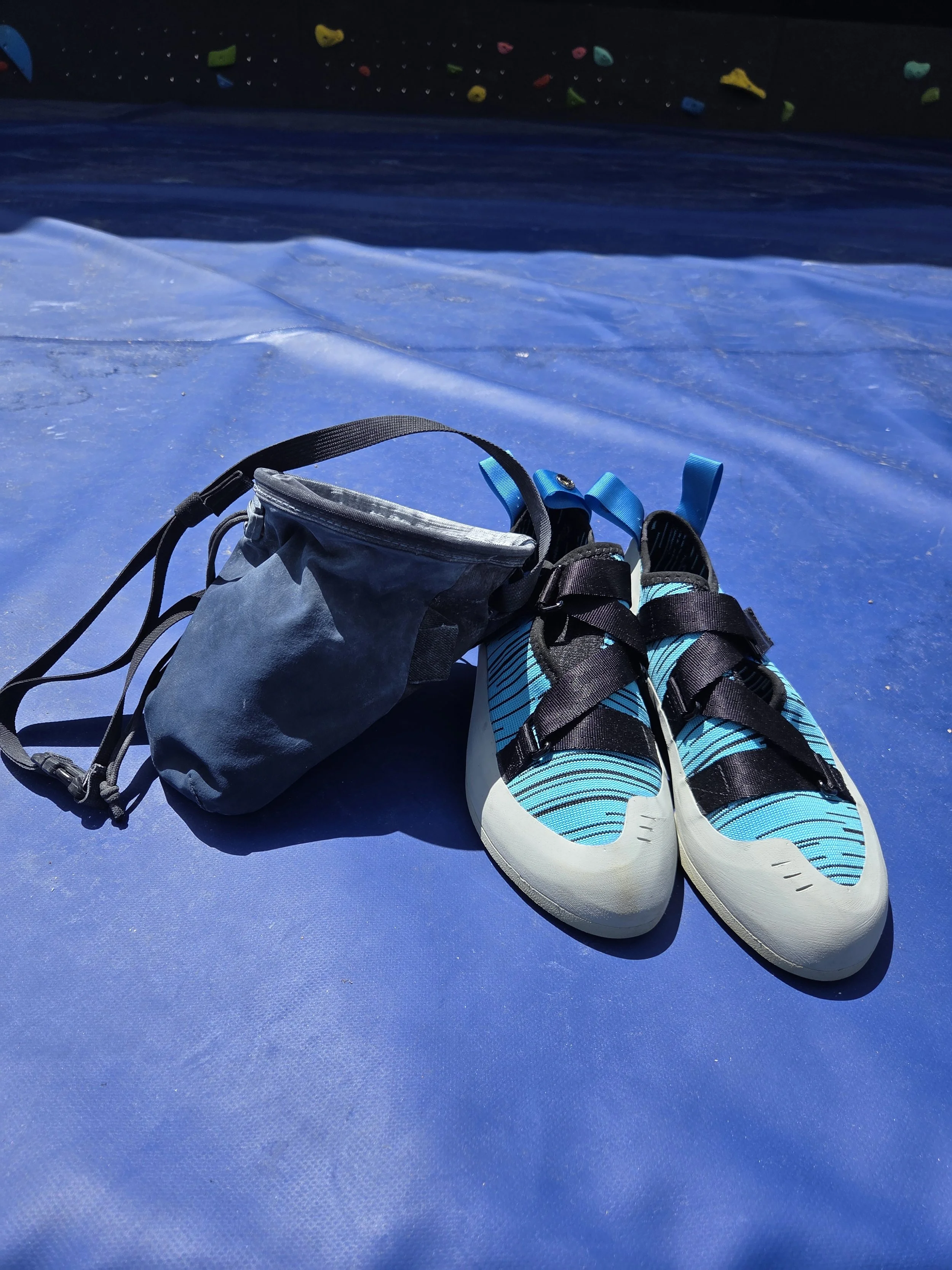 A pair of blue and white athletic shoes and a black backpack on a blue surface, with climbing holds visible on a wall in the background.