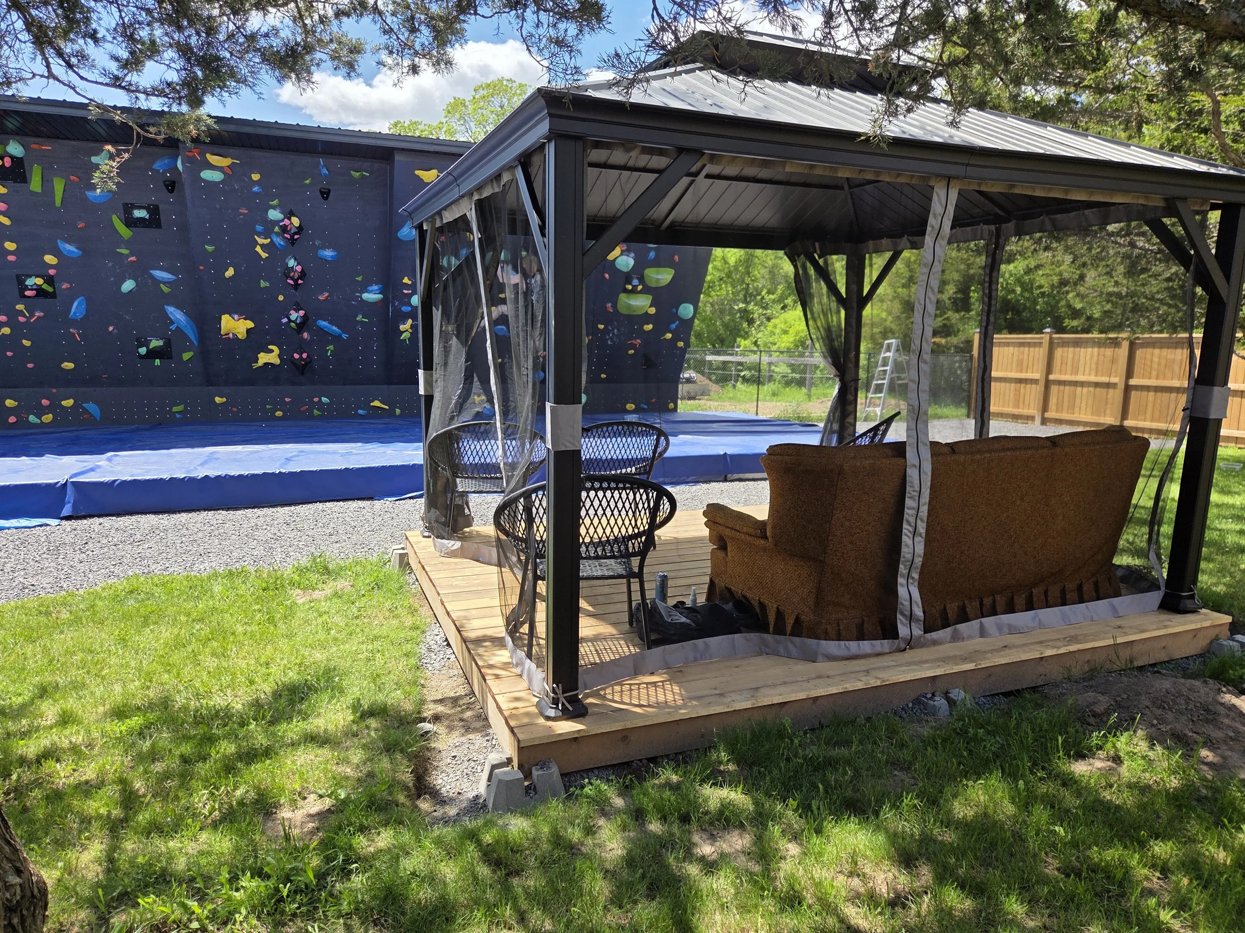 An outdoor scene showing a covered patio space with wooden decking, black metal supports, and transparent plastic curtains. Behind it, there is a blue climbing wall with colorful holds and a trampoline underneath it. The area is surrounded by green grass, trees, and a wooden fence, with a partly cloudy sky overhead.