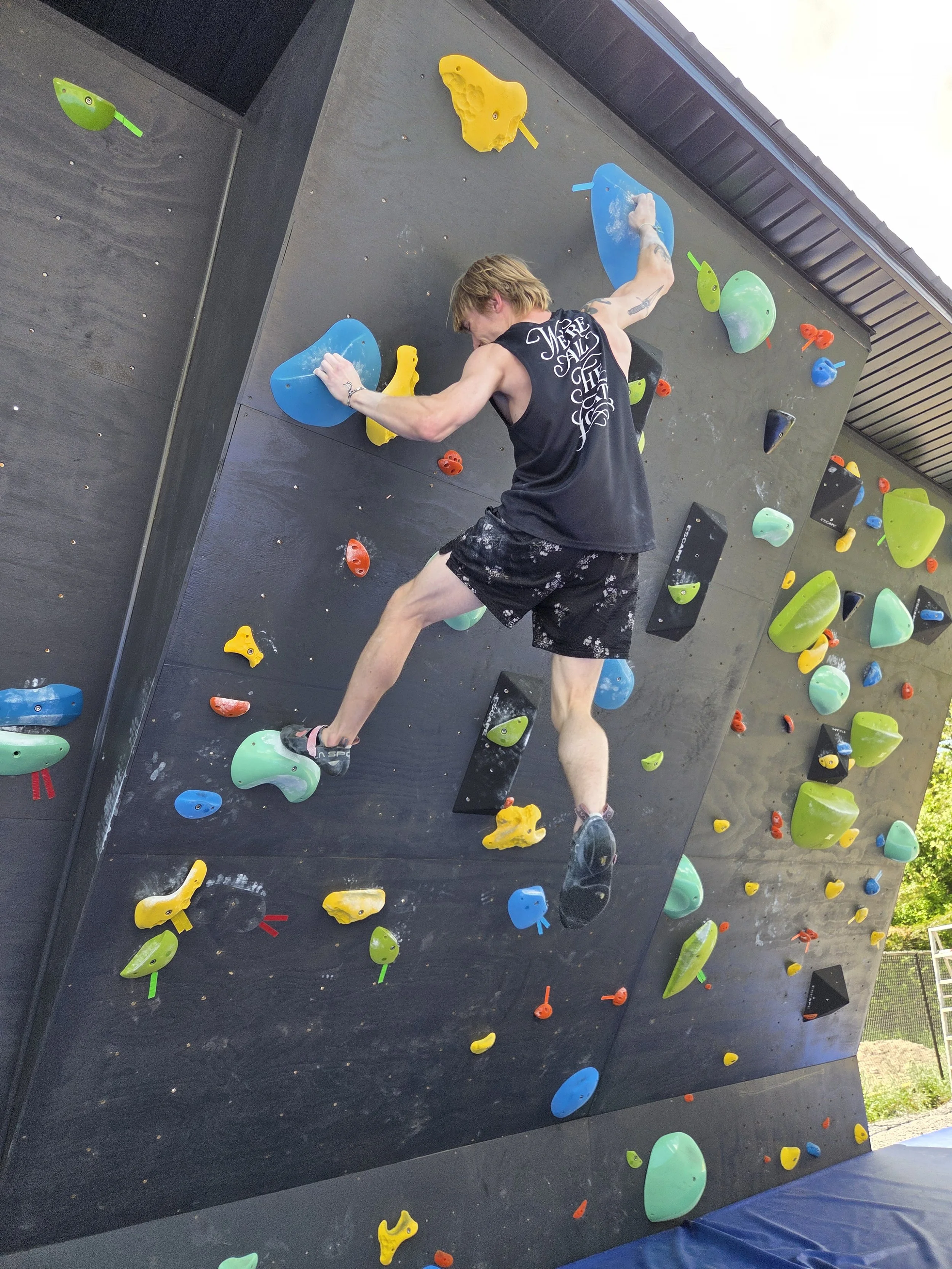 Person climbing an indoor rock climbing wall with various colorful holds, wearing a black tank top and black shorts.