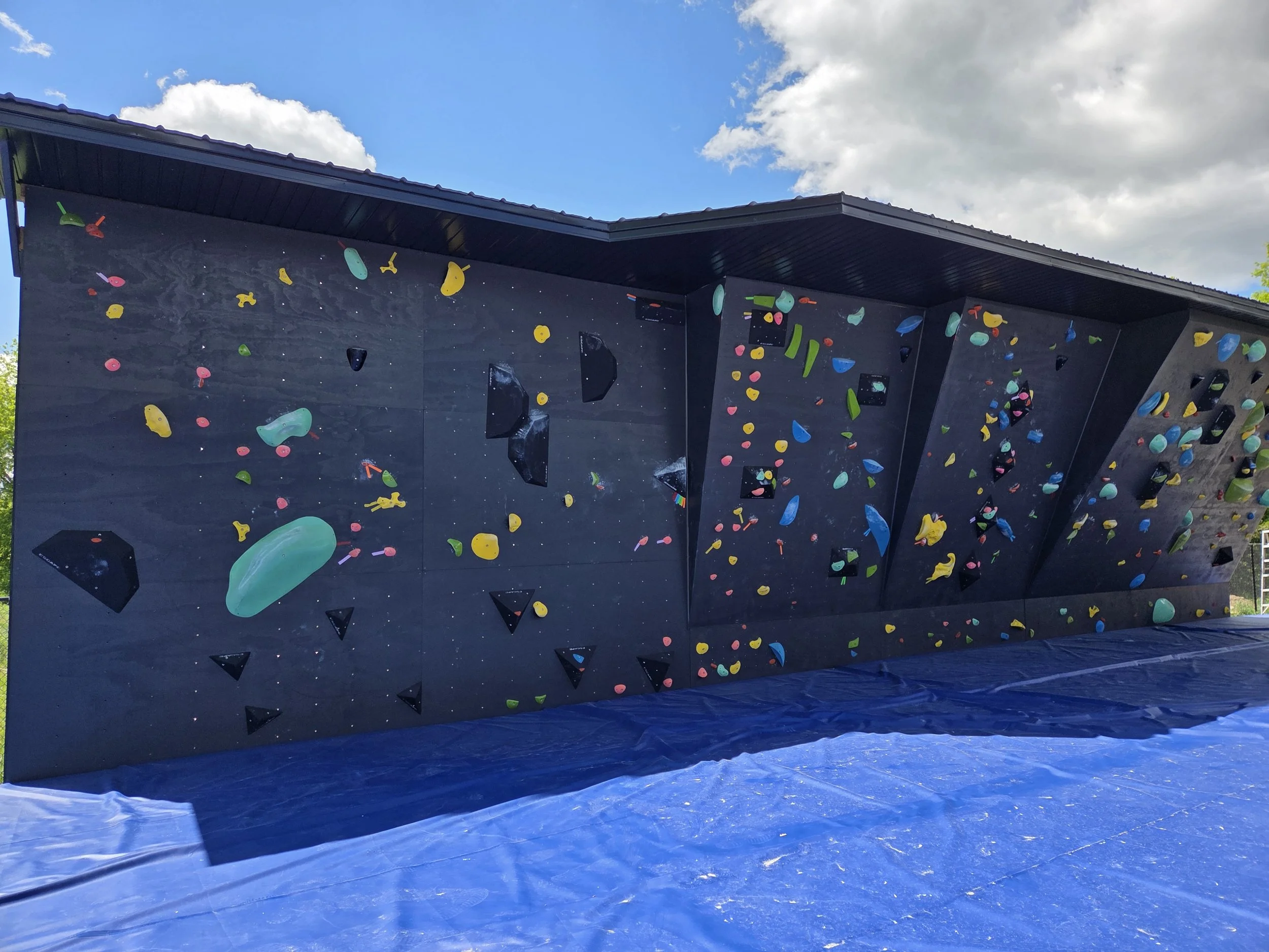 An outdoor rock climbing wall with colorful holds against a cloudy sky.
