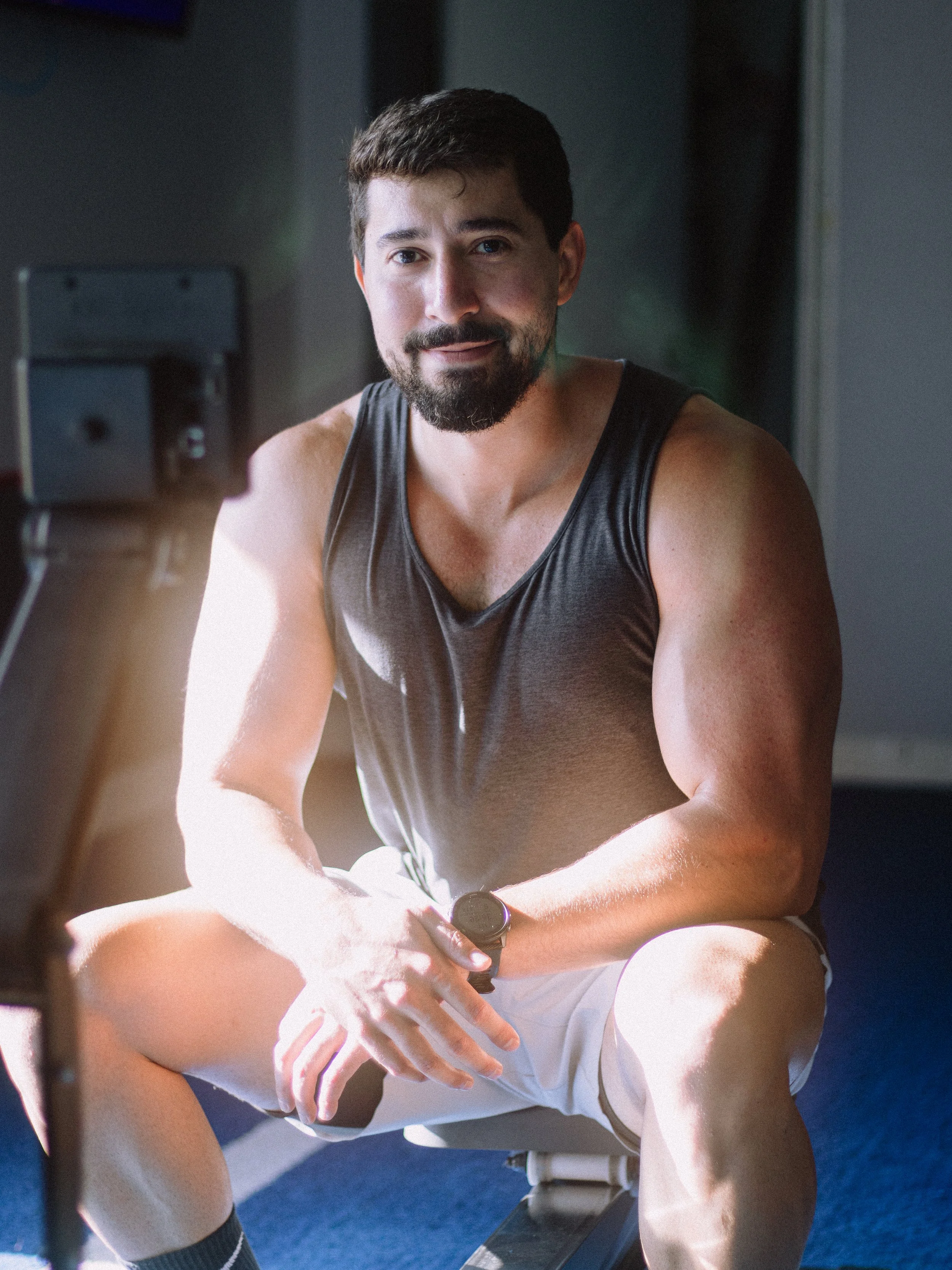 A man with a beard and mustache, wearing a sleeveless shirt and shorts, sitting on a bench or gym equipment, looking at the camera, with a smartwatch on his wrist.