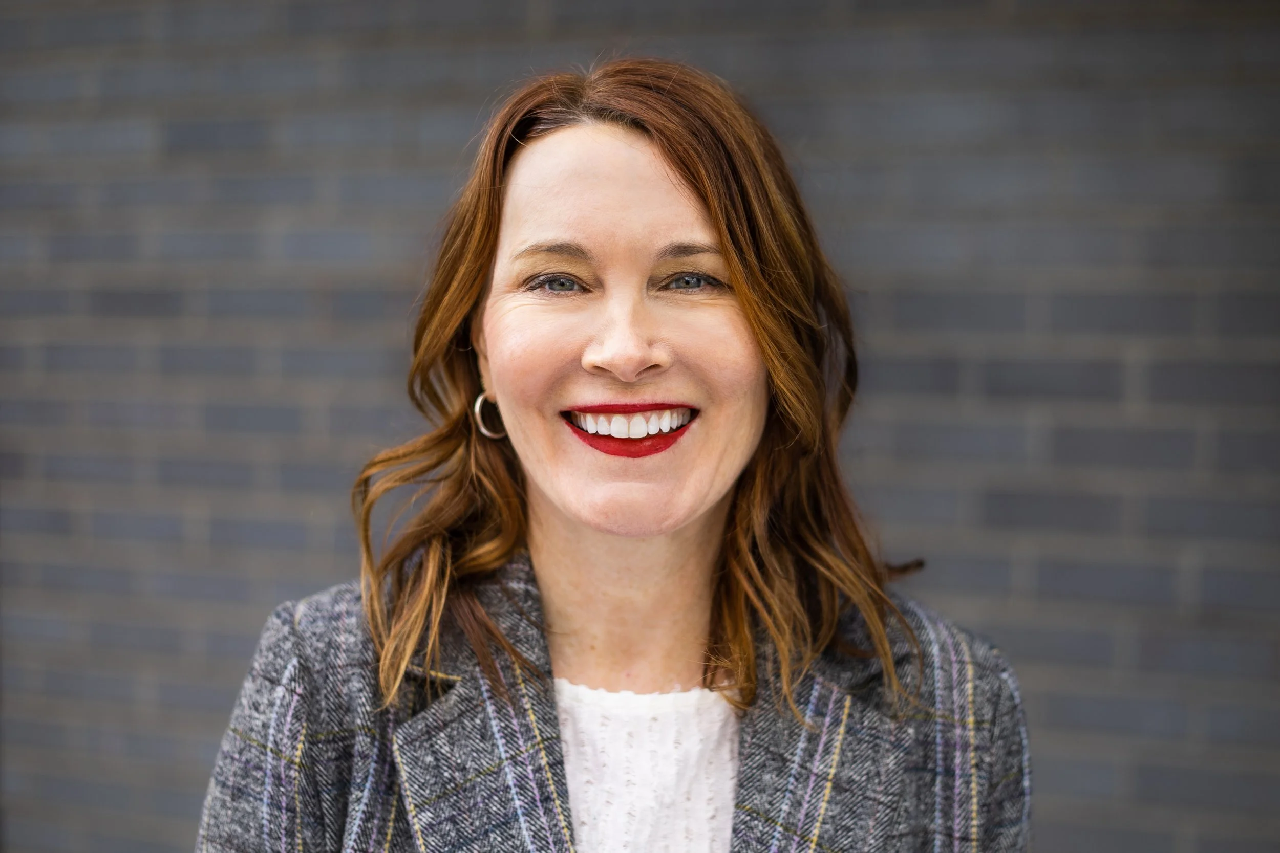 A woman with red hair and blue eyes smiling against a dark brick wall background.