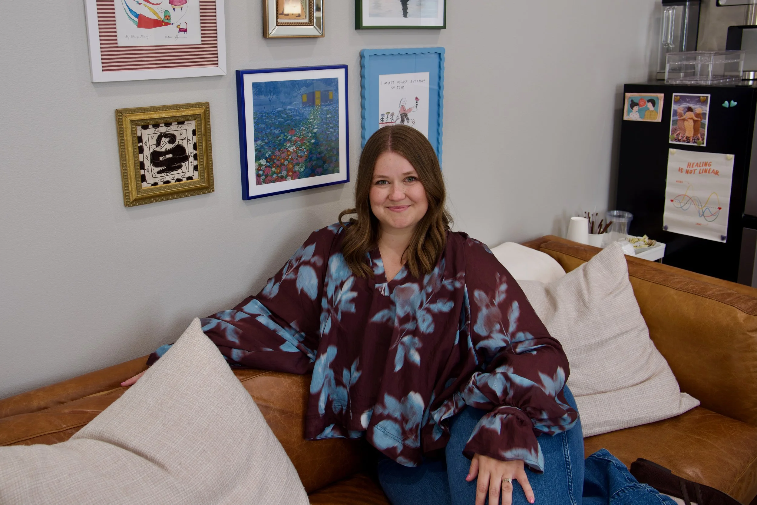 A woman with long brown hair sitting on a brown couch, smiling. She is wearing a purple and blue patterned top with jeans. Behind her, there is a wall decorated with framed pictures and artwork. To the right, part of a black piece of furniture with photos and a poster is visible.