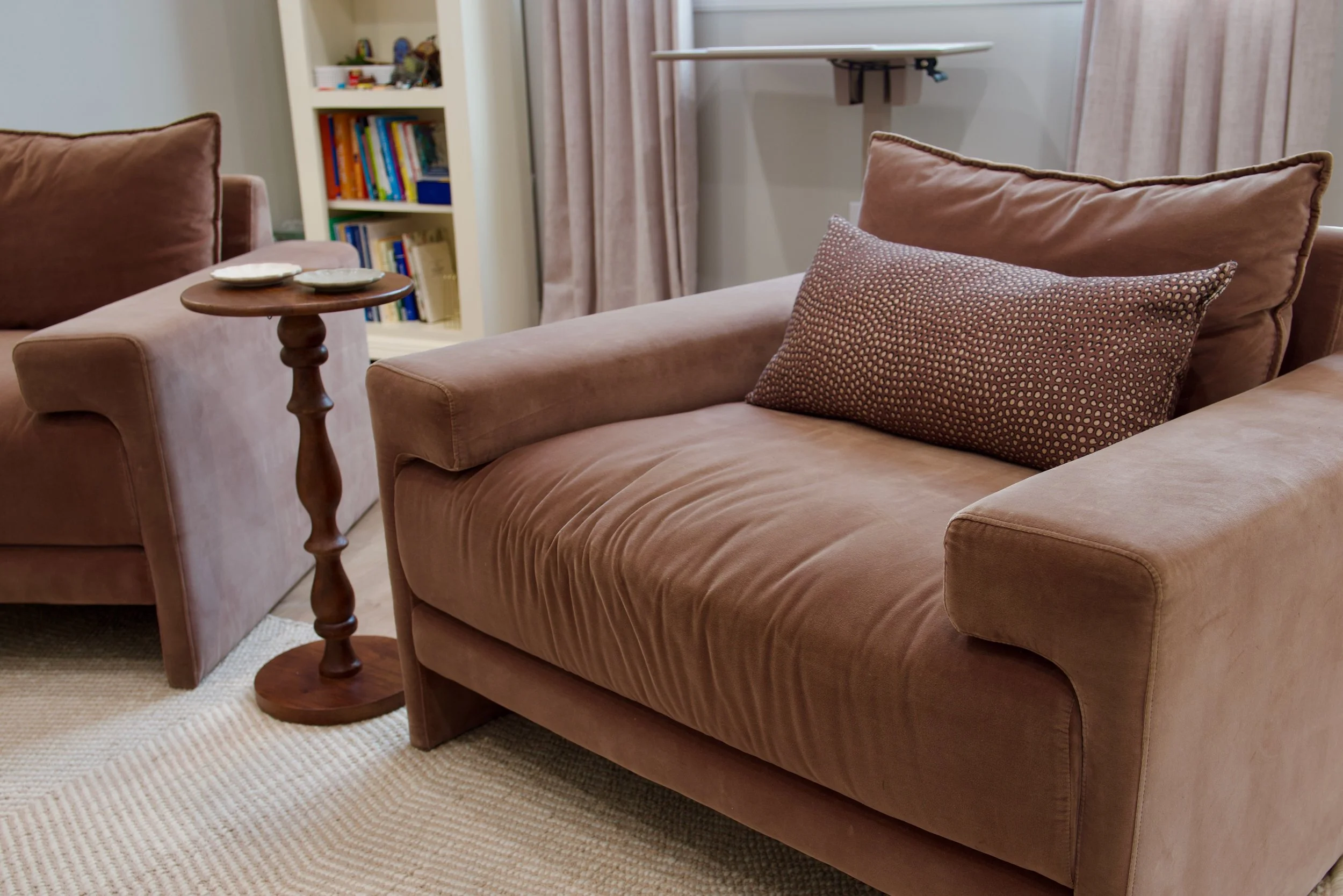 A living room with a pink sofa, large cushions, a wooden side table with coasters, a cream-colored carpet, a bookshelf with books, and light-colored curtains.