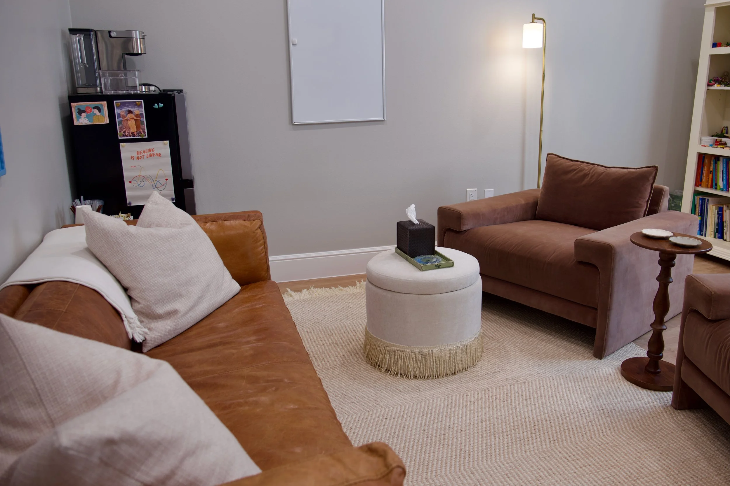 A cozy living room with beige carpet, brown and white sofas, a round beige ottoman, a wooden side table, a bookshelf filled with books, and a tall white lamp. There's a small black tissue box on the ottoman, a tissue protruding from it, and a white wall with an empty whiteboard.