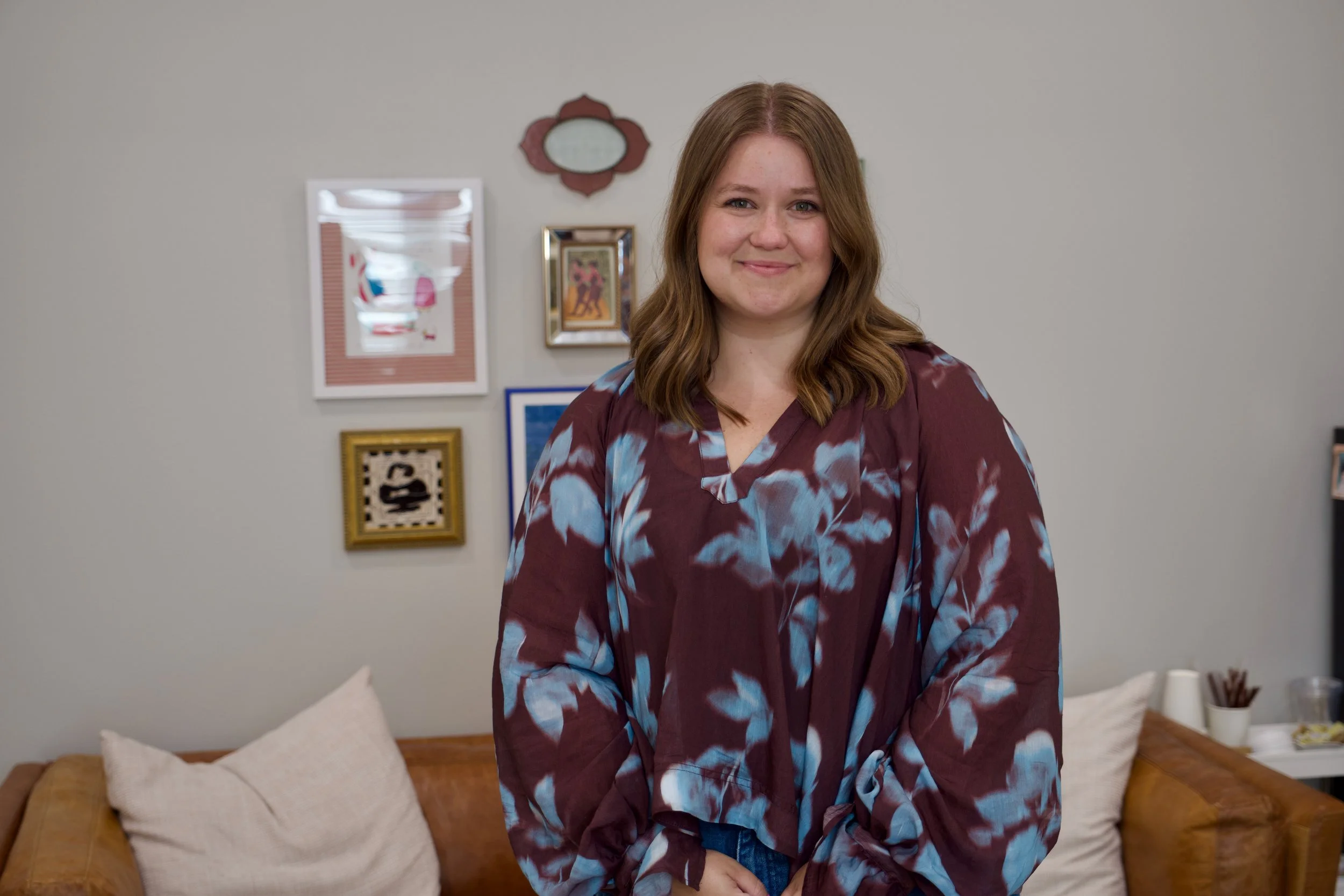 A woman with brown hair smiling into the camera, wearing a maroon, blue, and white patterned blouse, standing in a room with a wall decorated with framed pictures and a mirror.