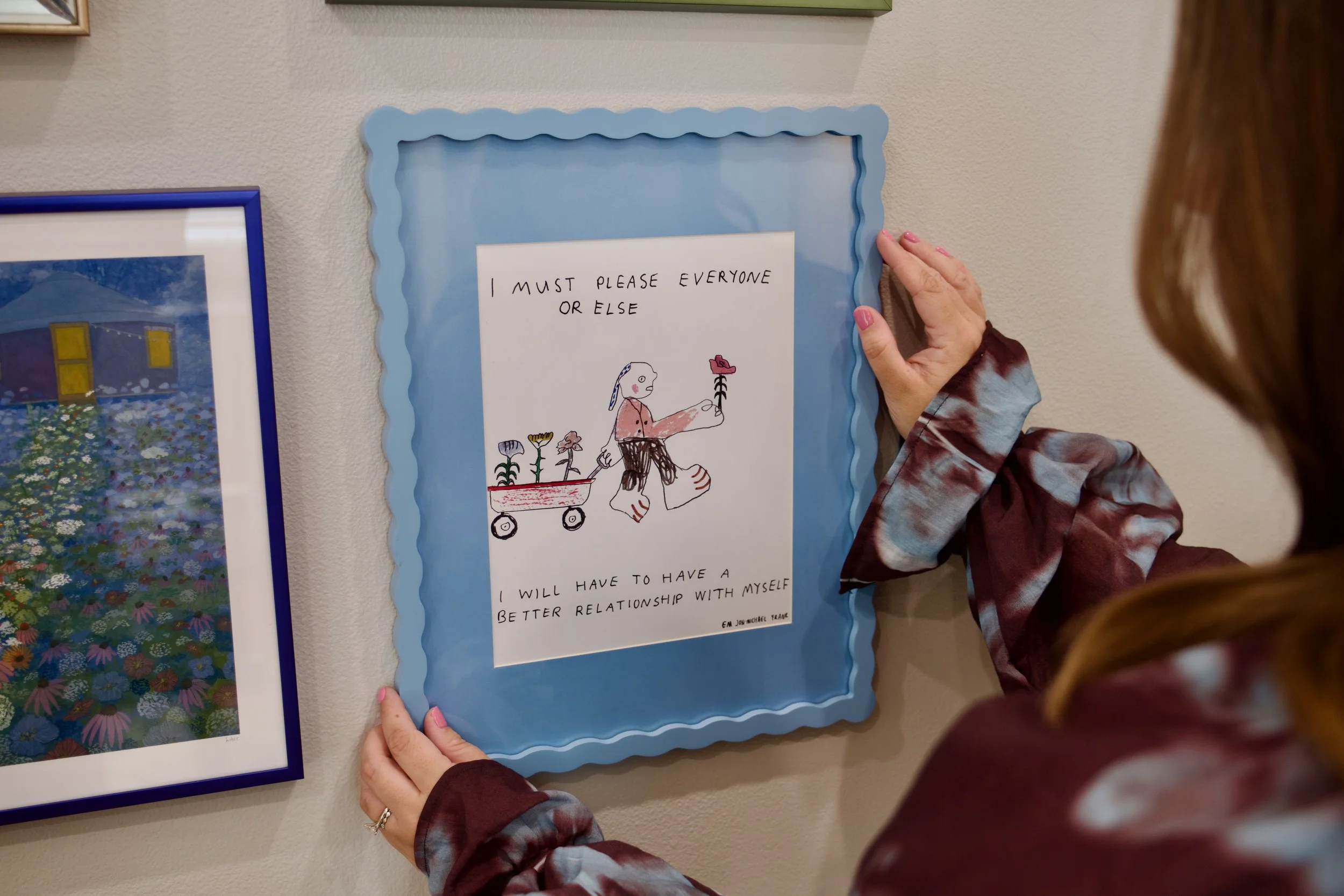 A person hanging an illustrated sign on a wall. The sign has the words 'I MUST PLEASE EVERYONE OR ELSE' and features a drawing of a character with a flower in hand, walking with a wagon of flowers.
