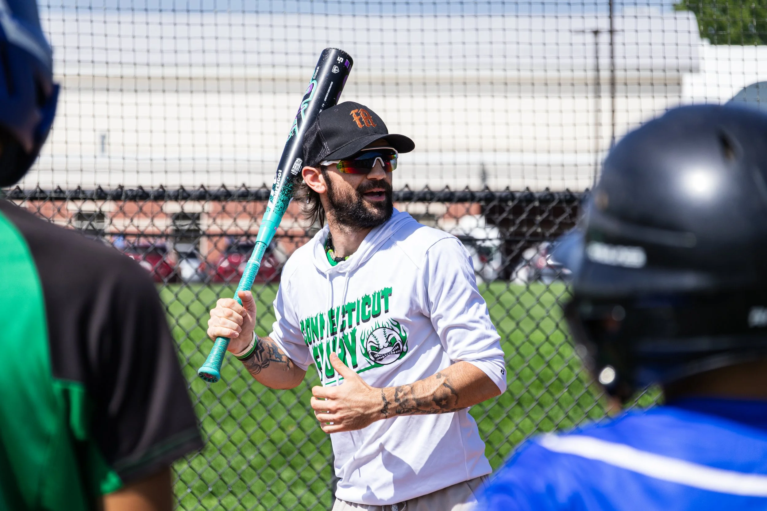A man with dark hair, a beard, tattoos, wearing sunglasses, a black cap, and a white T-shirt with green and black text, is holding a baseball bat and talking to people around him at a baseball field.