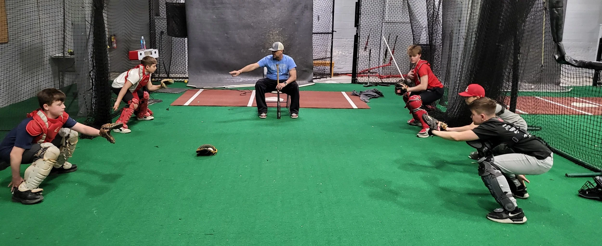 A youth baseball team practicing inside a batting cage with a coach giving instructions, all team members are in catching gear and squatting in ready positions.