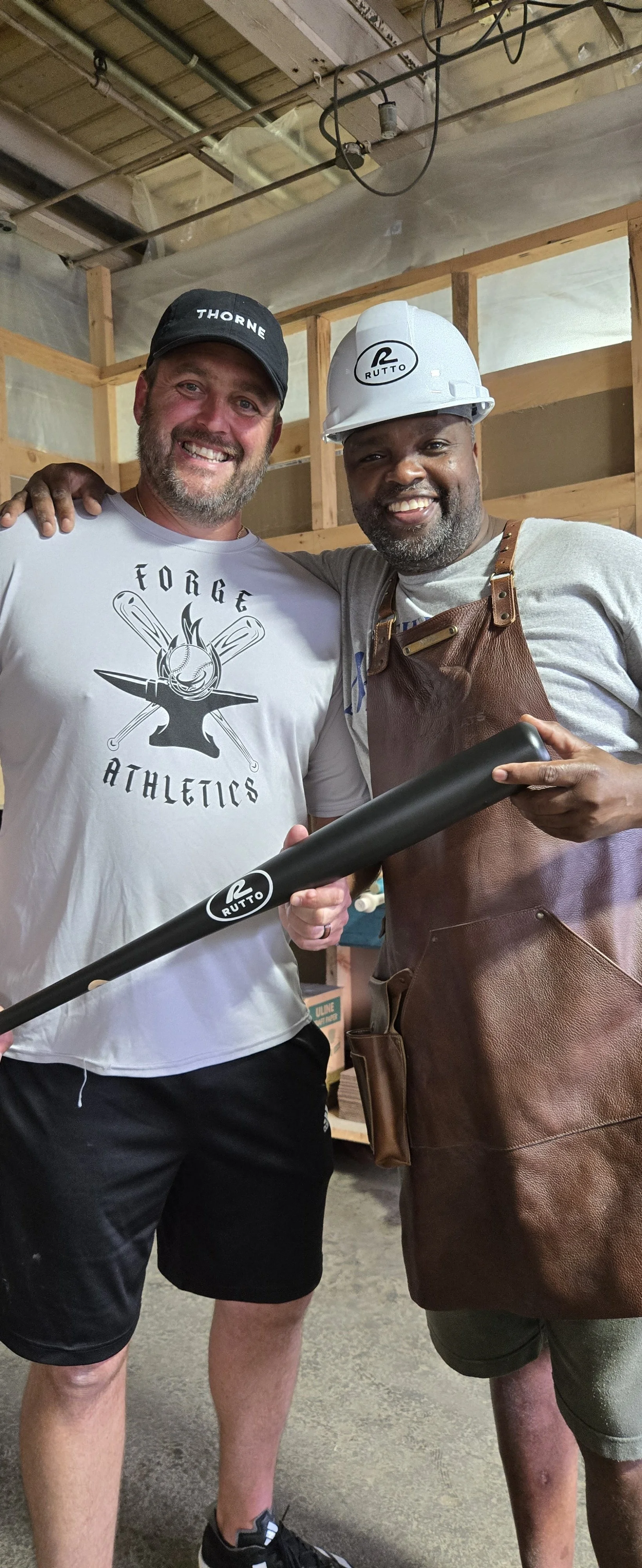 Two smiling men in casual and work attire holding a black baseball bat with the RUTTO logo at a construction site.