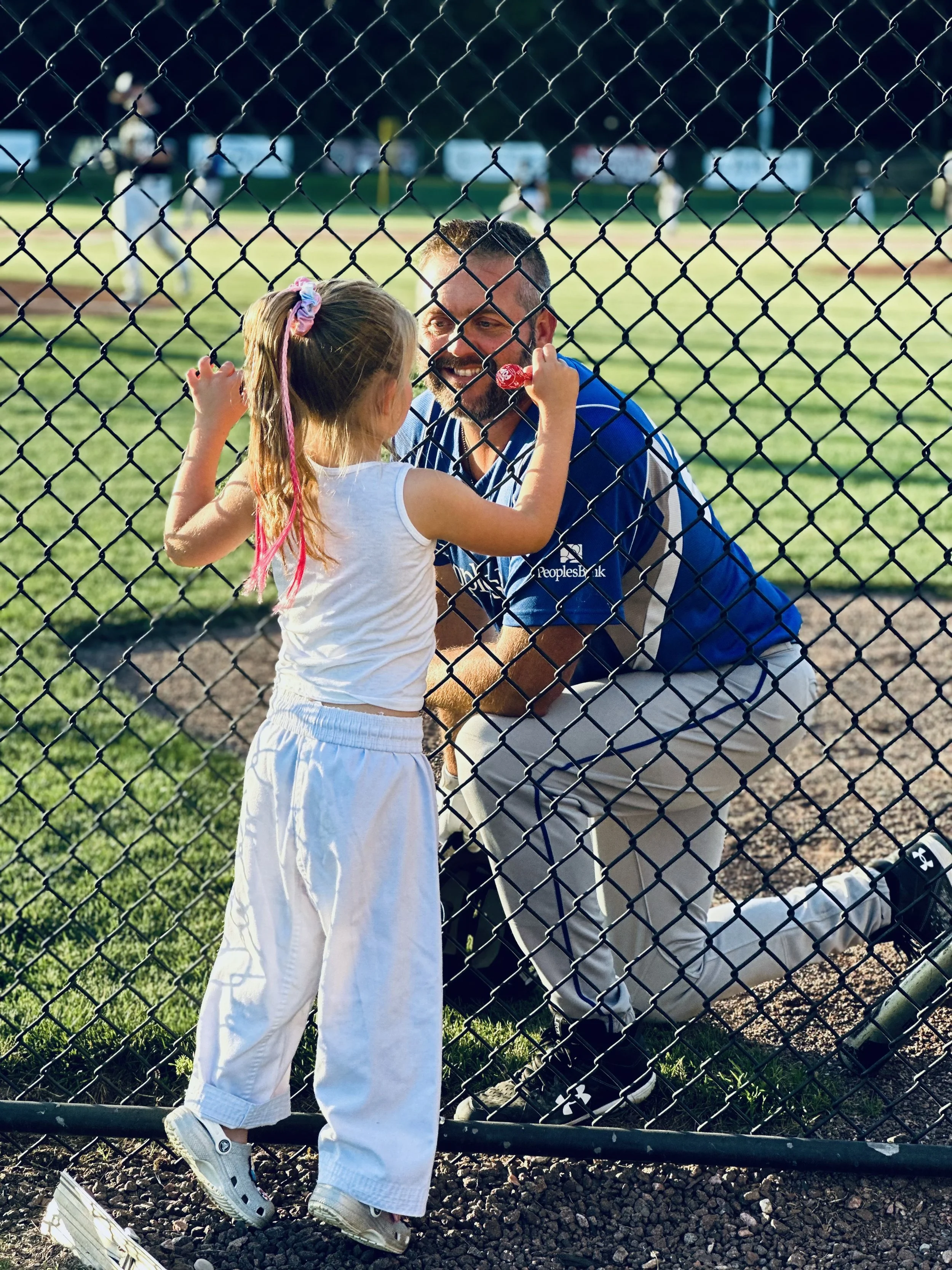 A young girl and a man on a baseball field behind a chain-link fence, smiling and talking, with other players in the background.