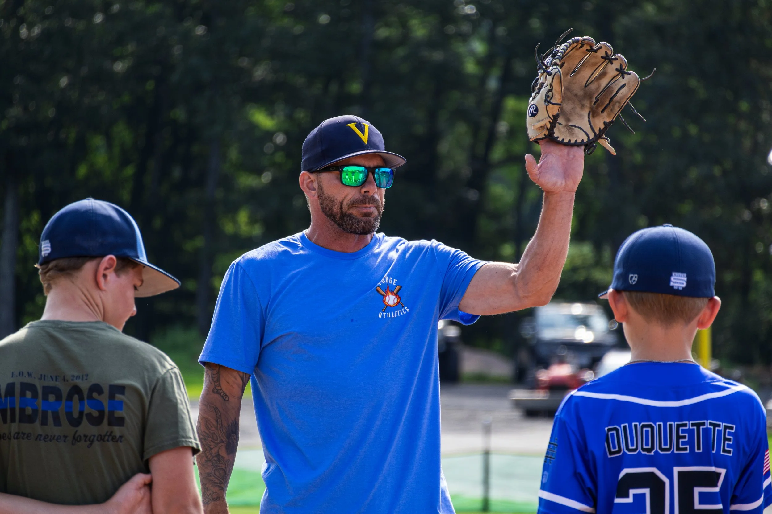 A group of children and a coach talking outdoors on a baseball field. The coach, a man with tattoos and wearing sunglasses and a navy cap with a yellow 'V', is raising a baseball glove. The children are wearing T-shirts and baseball jerseys.
