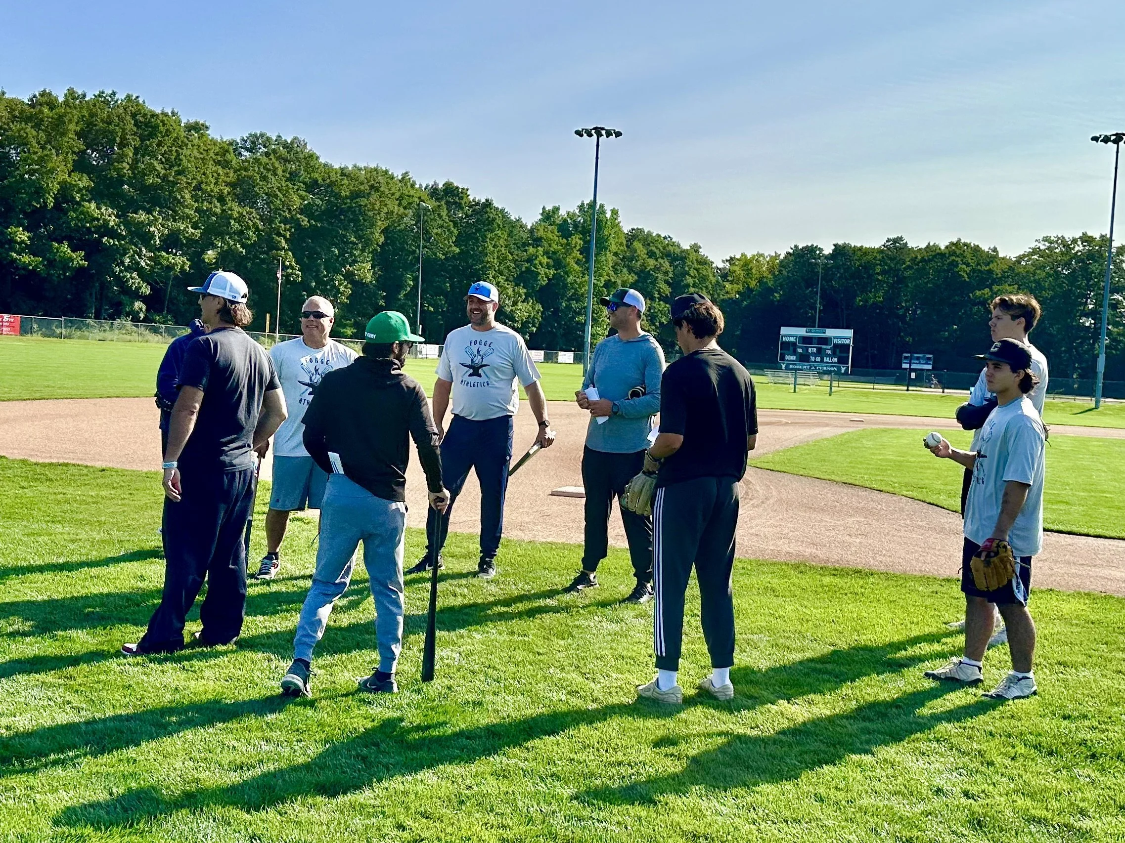 A group of young men and two older men standing on a baseball field grass, engaged in conversation, with some holding baseball equipment. They are dressed in athletic casual clothing, and the background features green trees, a scoreboard, and tall li