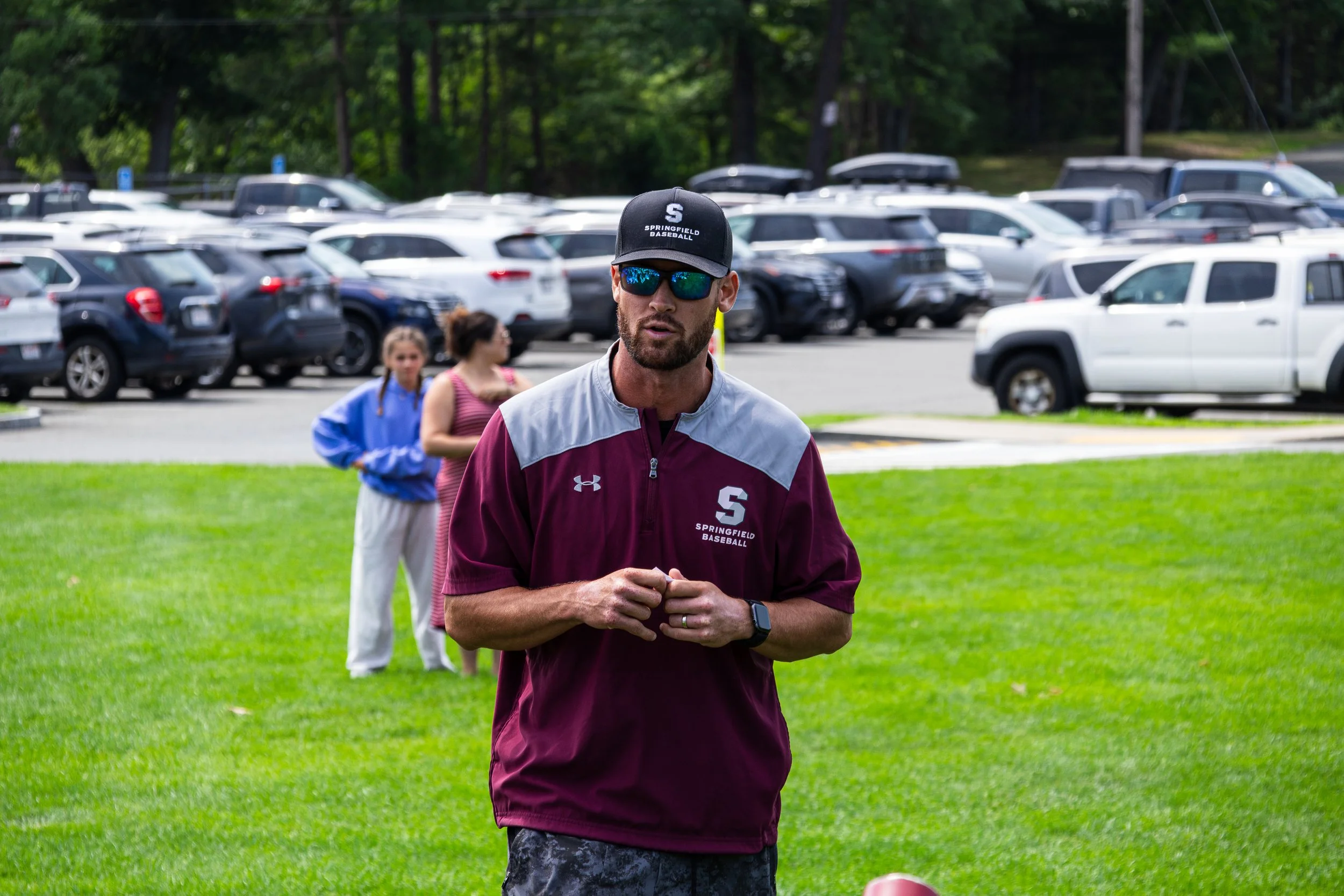 A man wearing sunglasses, a black cap with 'Springfield Baseball' written on it, and a maroon and gray jacket stands outdoors on a grassy area, with blurred people and a parking lot filled with cars in the background.