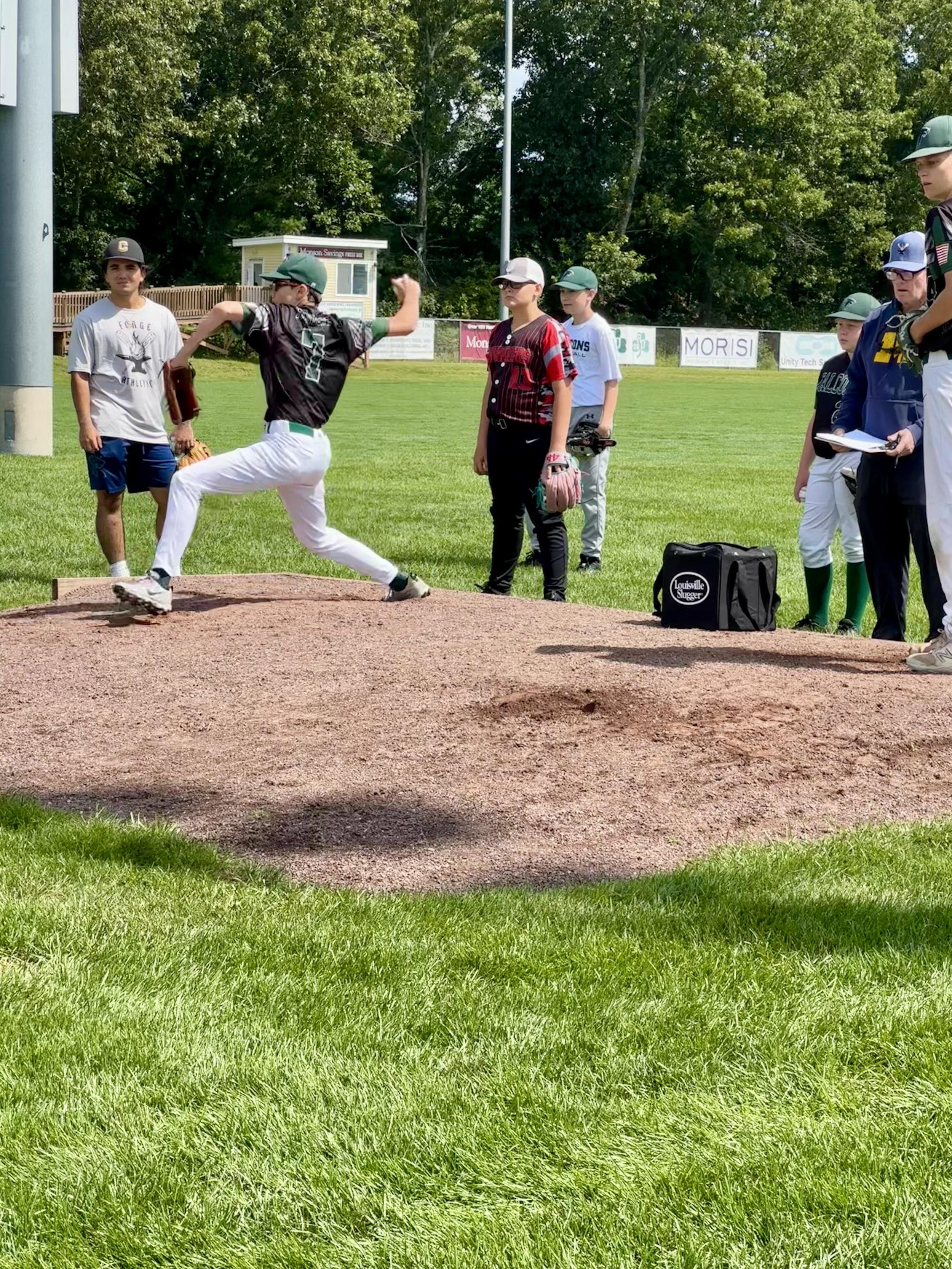 A youth baseball pitcher in black jersey and white pants throwing a pitch on a baseball field. Several spectators and players stand nearby, watching the game. The field is grassy and enclosed by a fence with trees in the background.