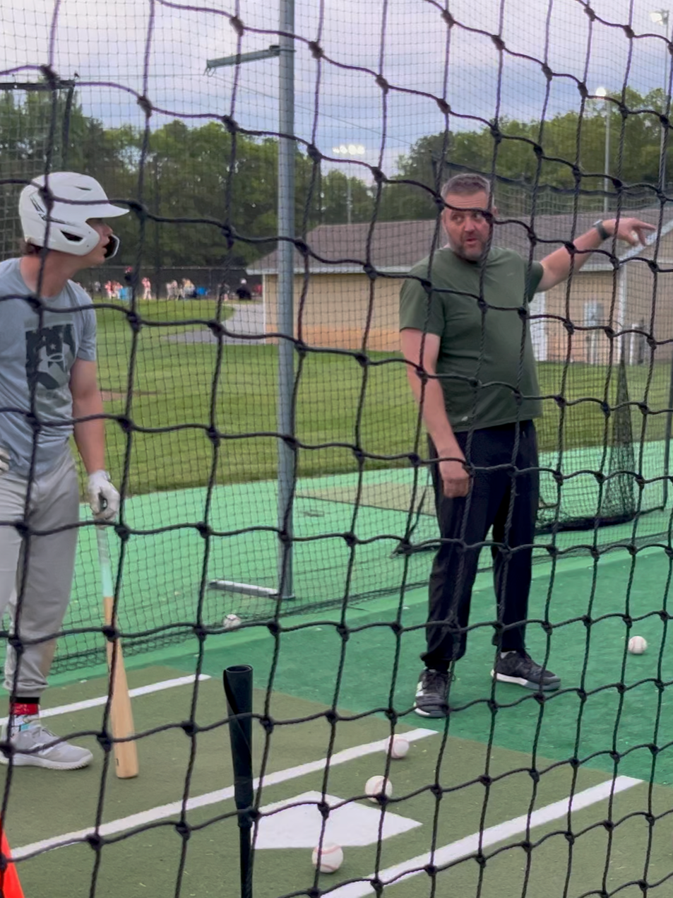 A coach giving batting instruction to a young baseball player at a batting cage, with the boy holding a bat and wearing a helmet, while the coach points and explains. Baseball balls are on the ground, and there is a net in front of them.