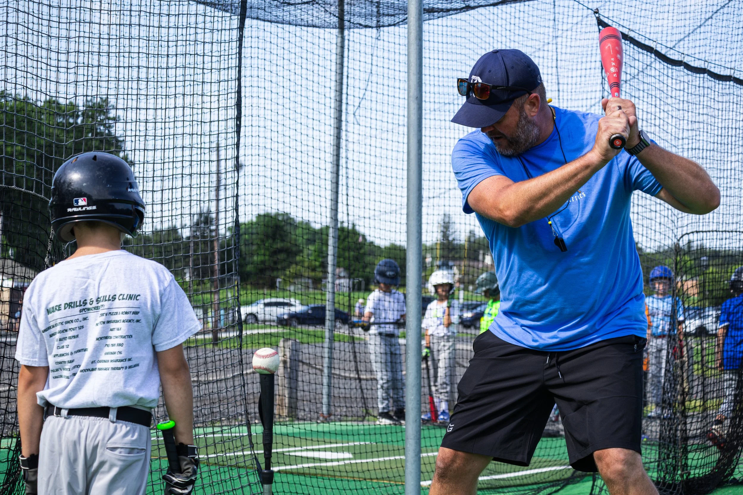 A baseball coach showing his batting stance to a young player inside a batting cage at a baseball field on a sunny day, with people and trees visible in the background.