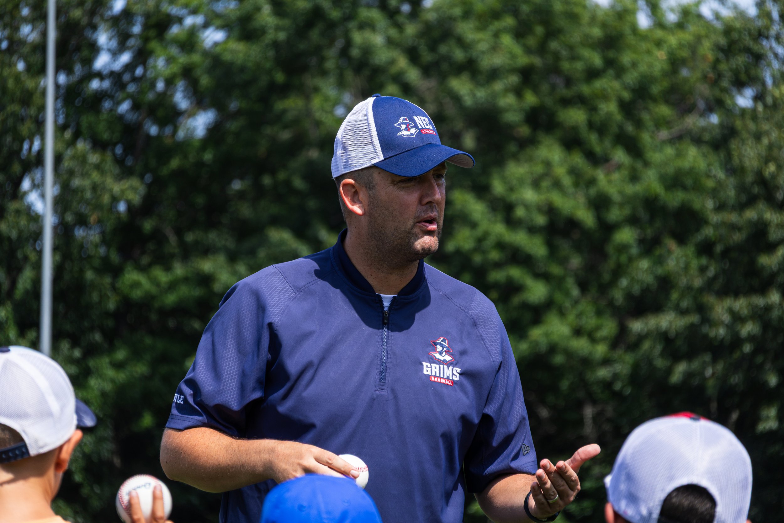 A man in a blue baseball uniform and cap with a logo, standing outdoors among children wearing white caps, holding baseballs, with green trees in the background.