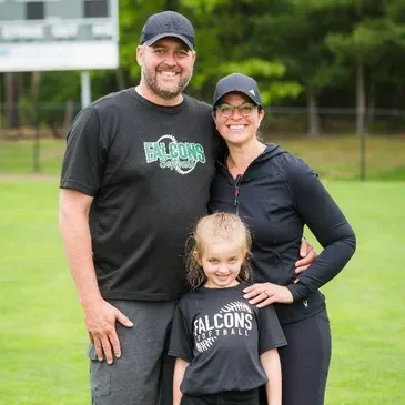 A family of three standing outdoors on a grassy field with trees in the background. The man and woman are smiling and wearing baseball caps, and the little girl is standing in front of them, also smiling.