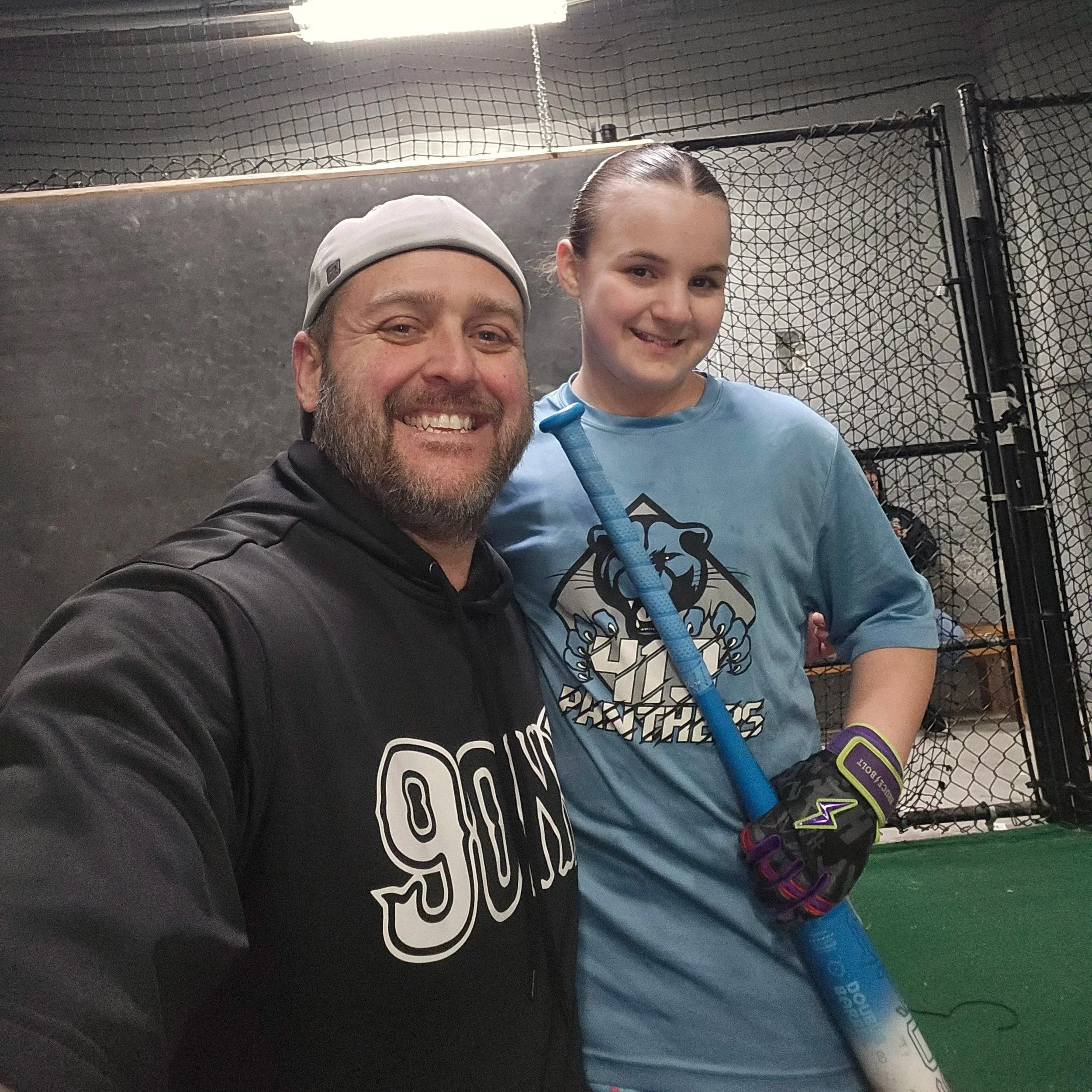 A smiling man and boy taking a selfie inside an indoor sports facility, with chain-link fencing and a grey wall in the background. The man is wearing a grey cap and a black jacket, and the boy is holding a hockey stick and wearing a light blue shirt.
