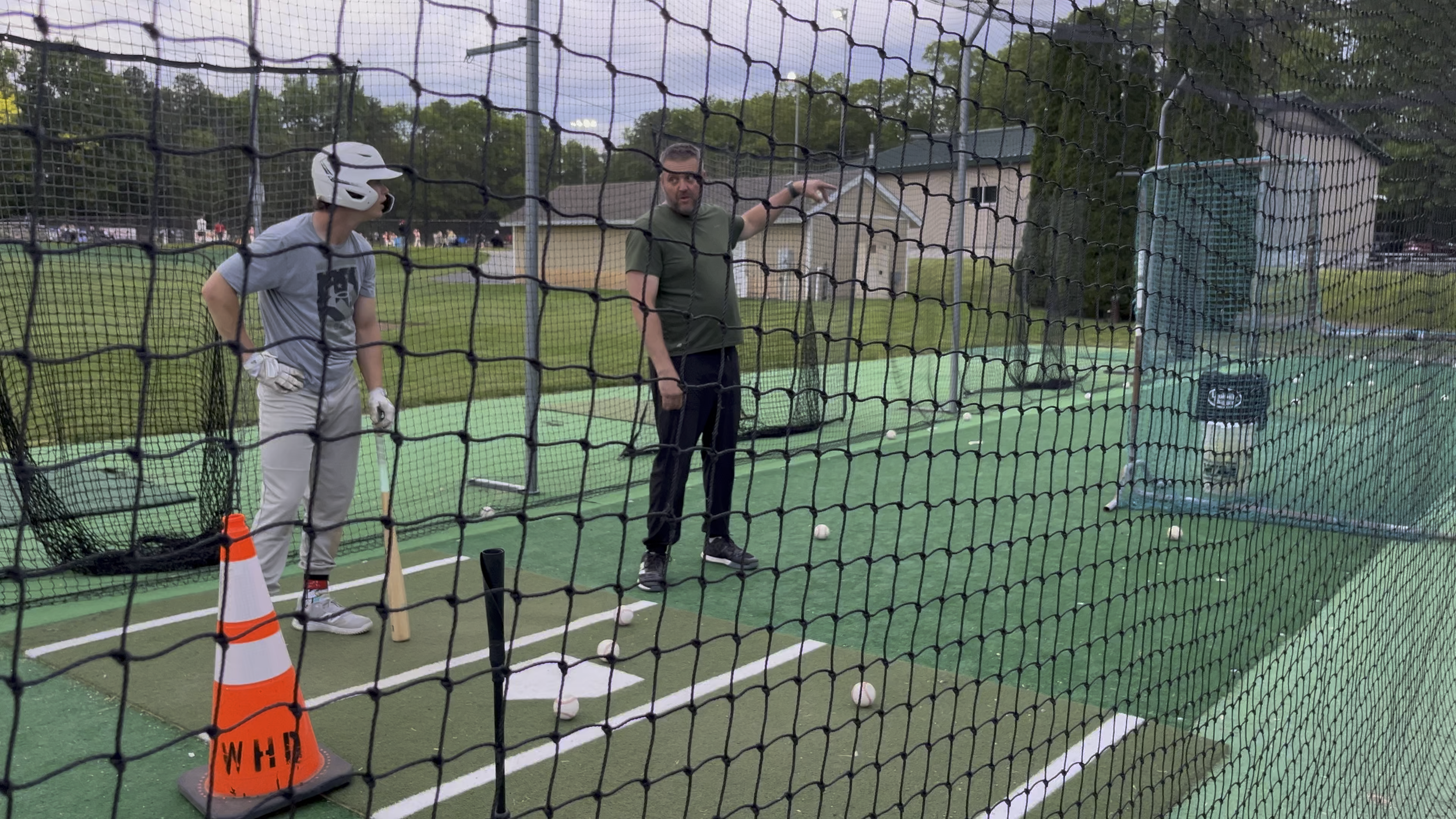 A man and a young person standing in front of a batting cage at a baseball facility, with a green turf surface, baseballs, sports equipment, and a building in the background, viewed through a safety net.