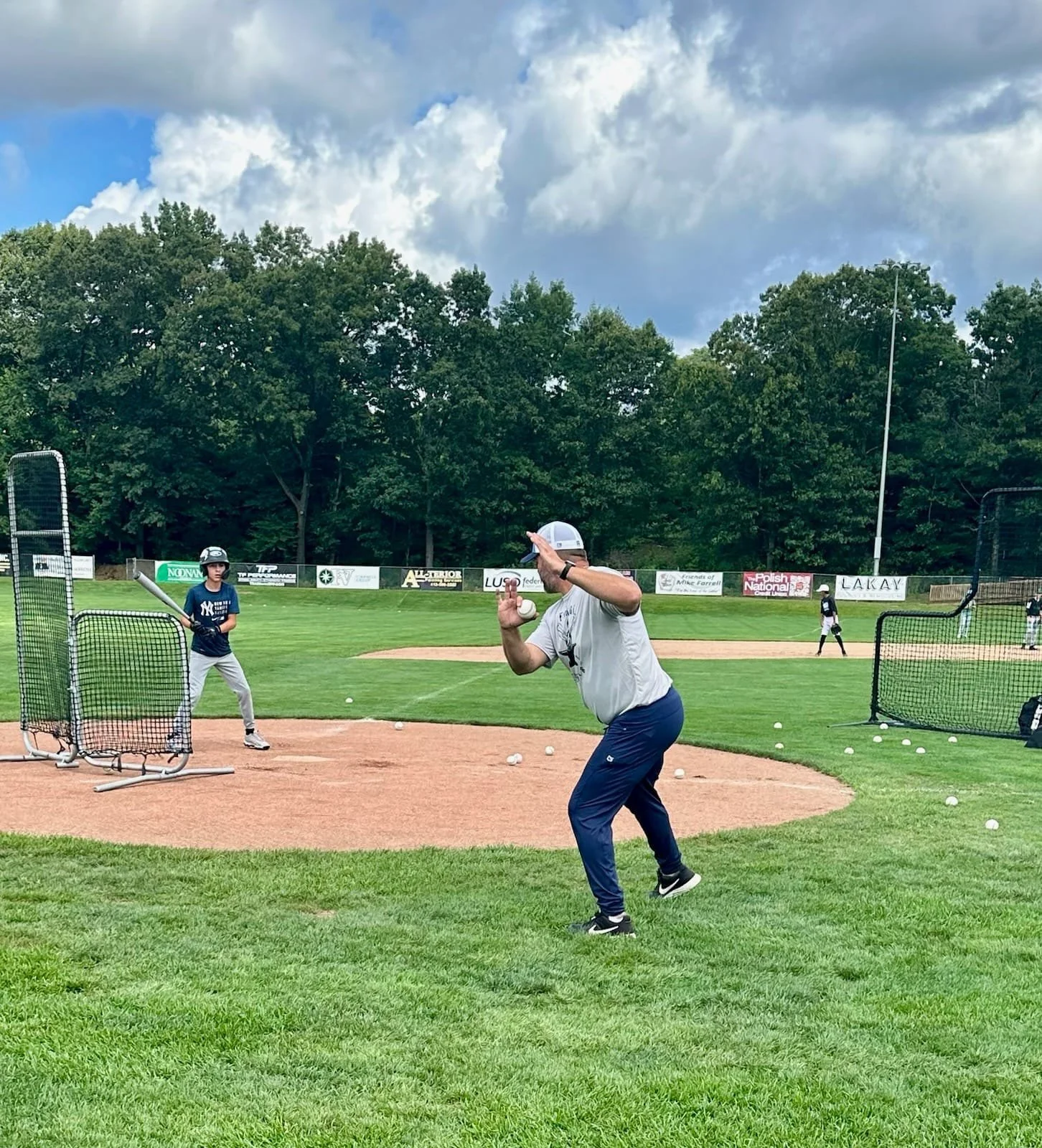 A baseball coach practicing batting with young players on a baseball field during daytime. Coaches and players are in the batting cage and around the field. The sky is partly cloudy with green trees in the background.