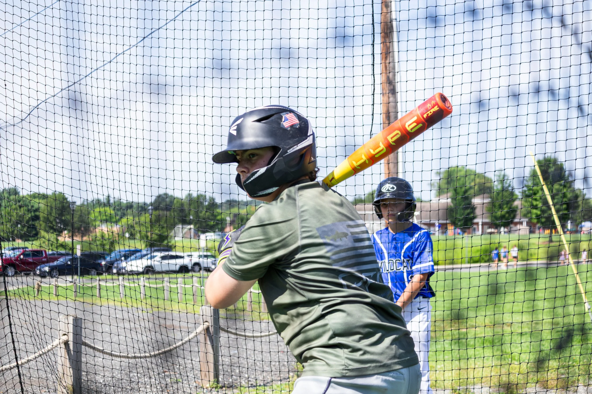 A young baseball player with a batting helmet practicing batting in the batting cage, with a girl in the background wearing a blue sports jersey and several parked cars visible behind a chain-link fence on a sunny day.
