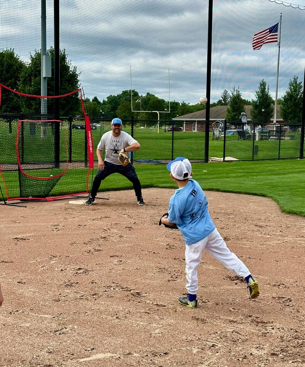 A boy in a baseball uniform preparing to catch a ball on a baseball field with a coach behind him near the batting cage, with an American flag flying in the background.