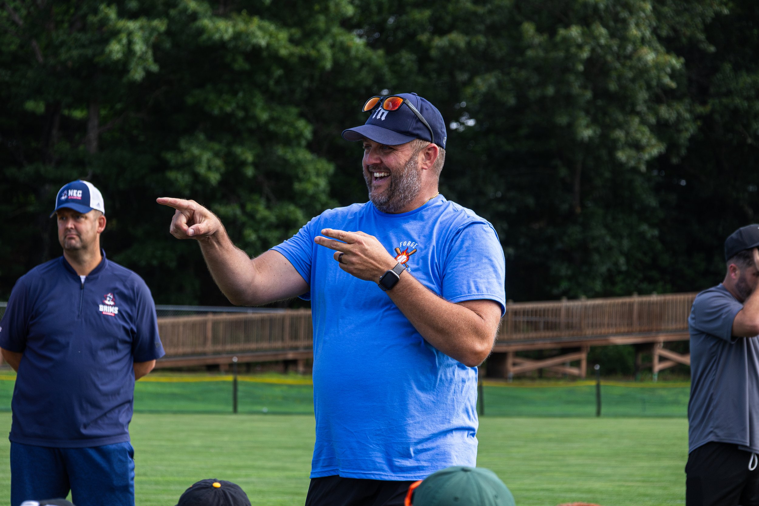 A group of men standing outdoors on a sports field, with one man in a blue shirt and cap smiling and pointing, others looking on, and a wooded background.