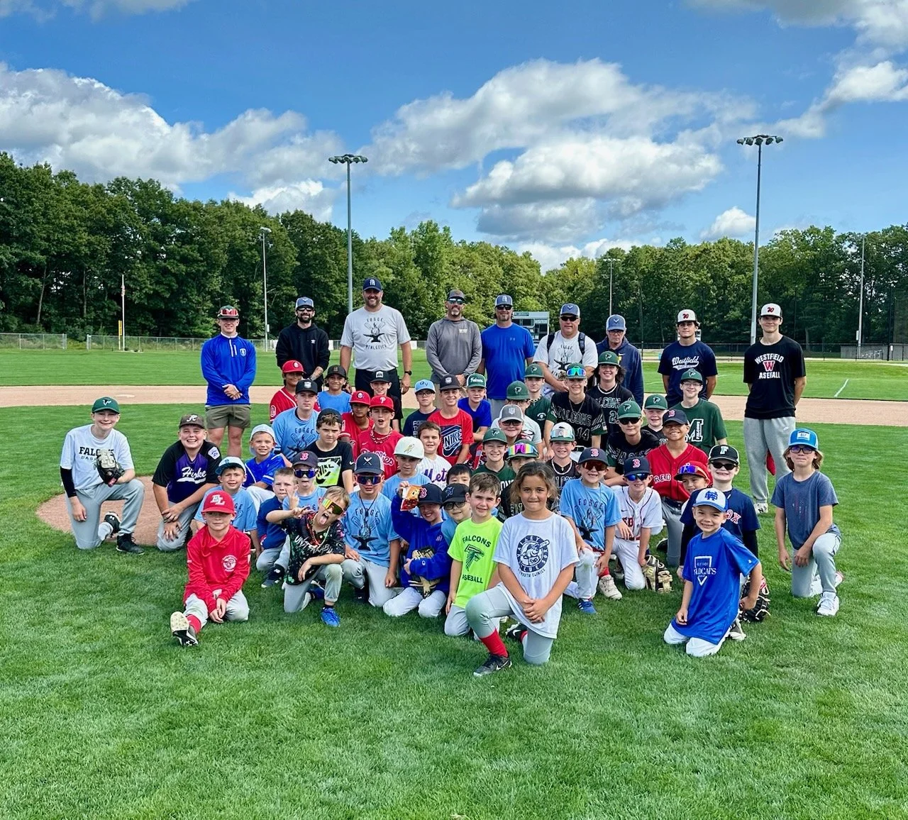 Group of children and coaches on a baseball field, posing for a team photo on a sunny day.