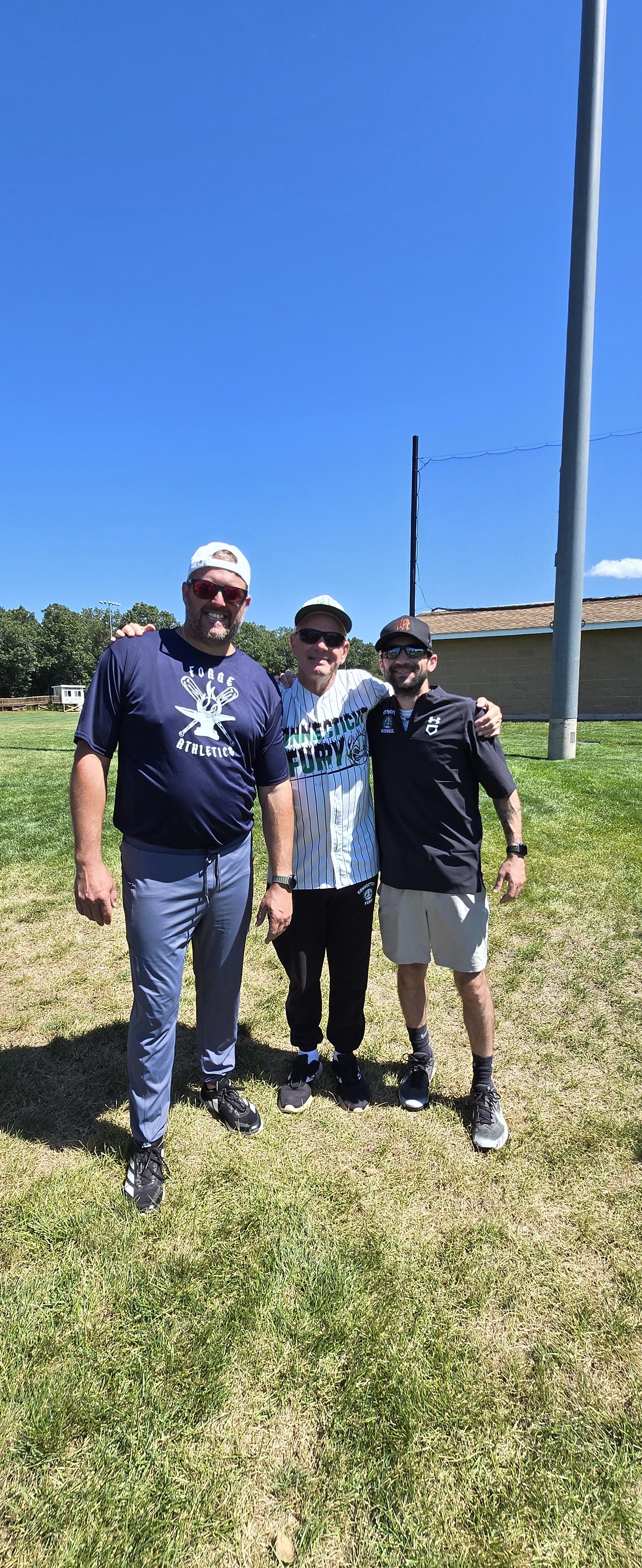 Three men standing on grass field, smiling, with clear blue sky overhead, a building, and a tall light pole behind them.