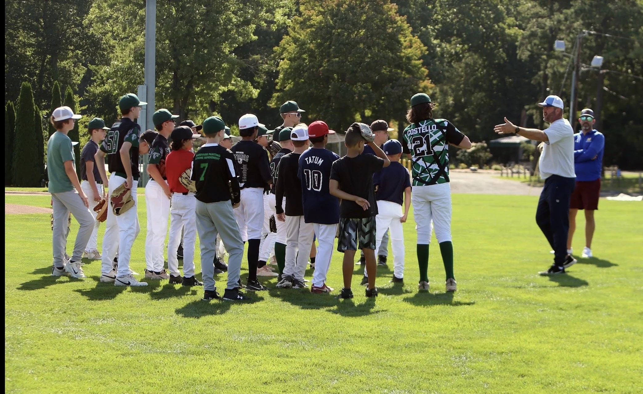 Youth baseball team listening to coach during practice on a sunny field with trees in the background.