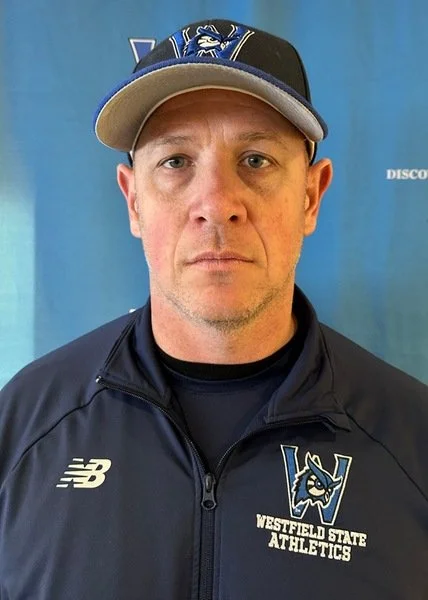 A man wearing a baseball cap with a Westfield State athletics logo and a windbreaker jacket with the same logo, standing in front of a blue background.