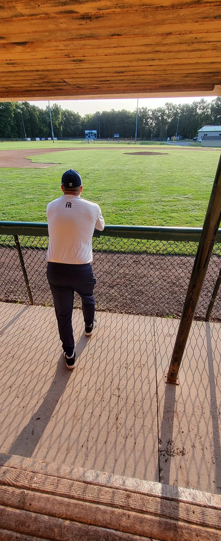 A man in a white shirt, dark pants, and a baseball cap stands on a dirt pathway, leaning on the fence and looking at a baseball field with green grass and brown dirt areas in the background.