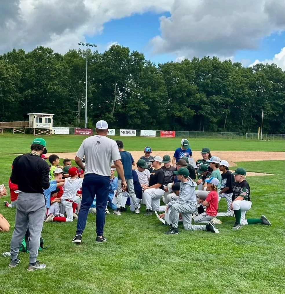 A group of youth baseball players and coaches gathered on a baseball field during daytime, with a grassy outfield, trees, and a cloudy sky in the background.