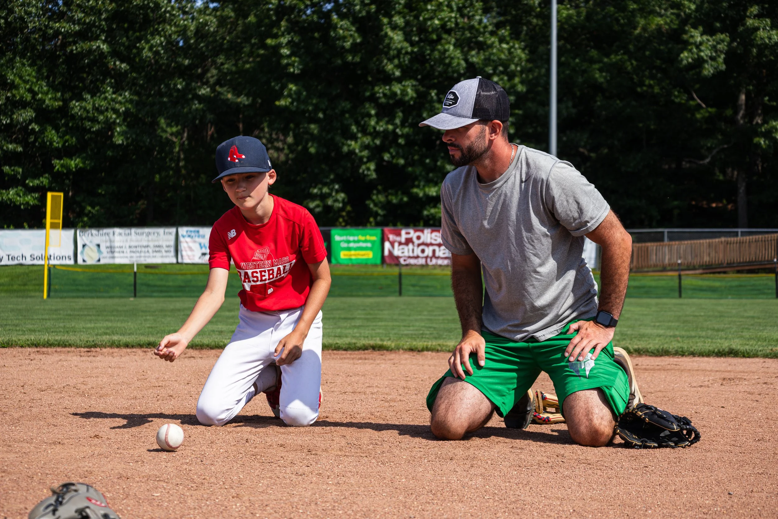 A young baseball player and an adult coach kneeling on a baseball field, with a baseball on the ground nearby. The young player is wearing a red shirt and white pants, and the coach is wearing a gray shirt, green shorts, and a black and gray cap. The