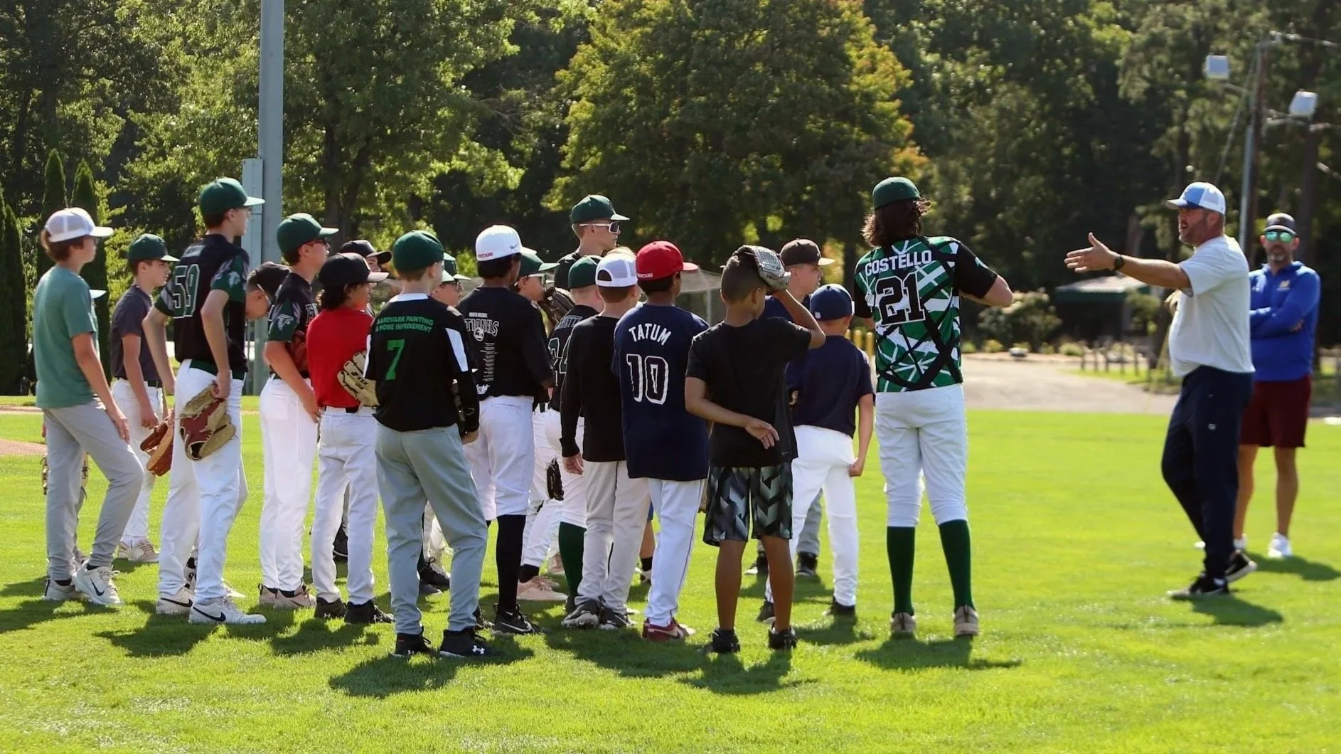 A group of young boys in baseball uniforms and hats stand on a grassy field listening to two coaches, one male and one female, giving instructions during a practice or game.