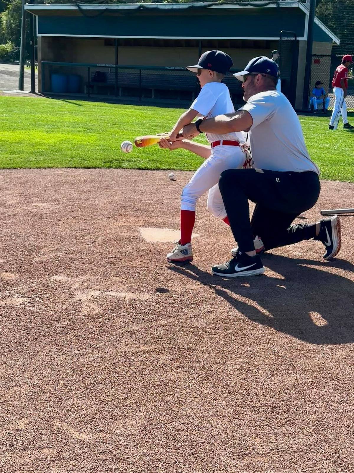 A young baseball player in a white uniform and red socks practicing batting with an adult coach kneeling in front of him, on a baseball field during daytime.