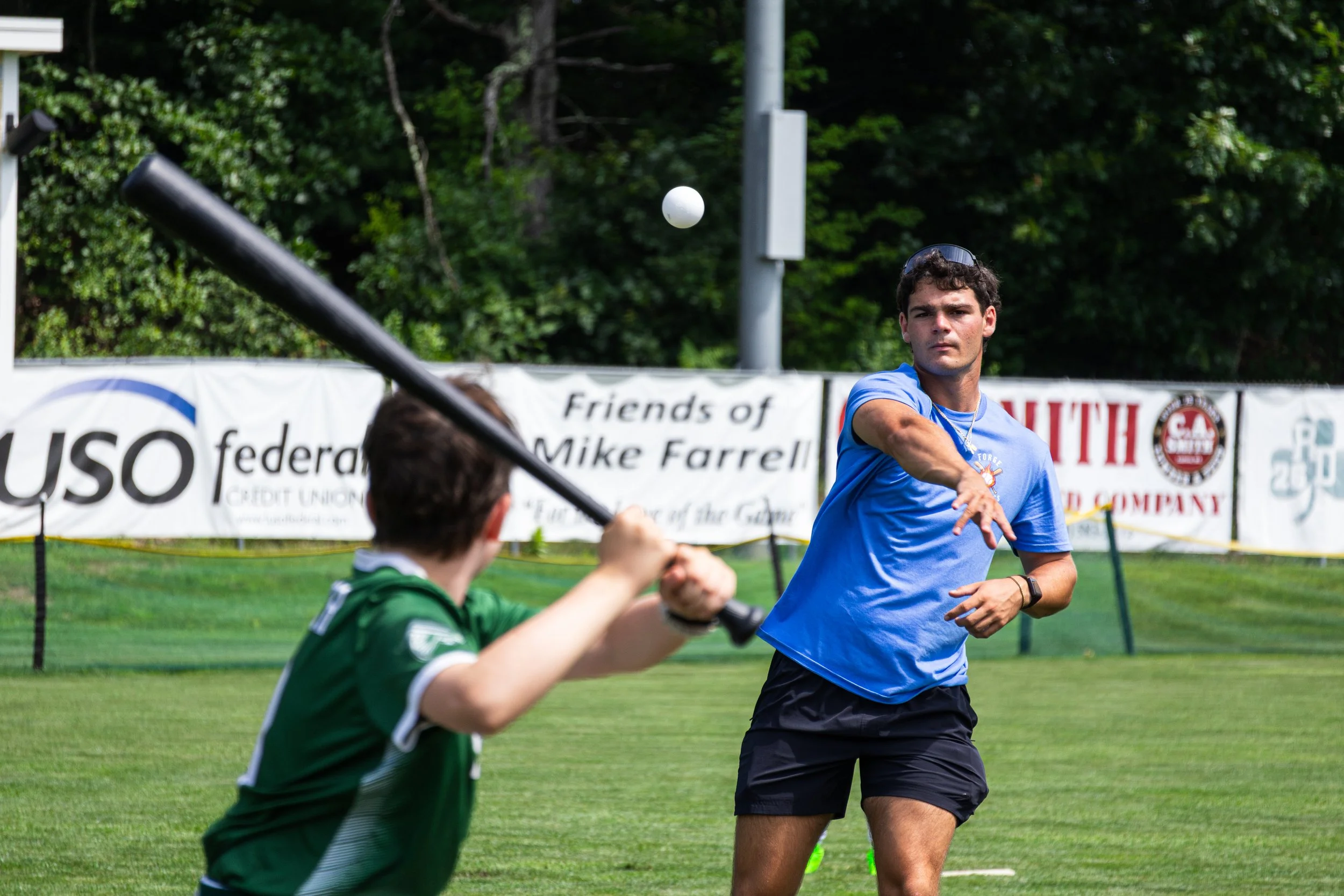 A young man in a blue athletic shirt and black shorts catching a baseball while another player in a green jersey throws the ball at a baseball field with a fence and trees in the background.