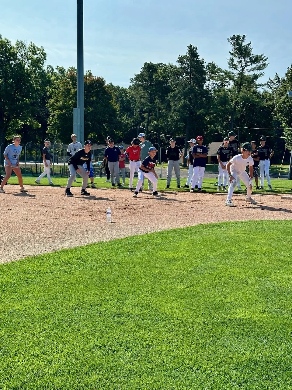Kids participating in a youth baseball practice or game on a baseball field with supporting players on the sidelines, with trees and utility poles in the background.