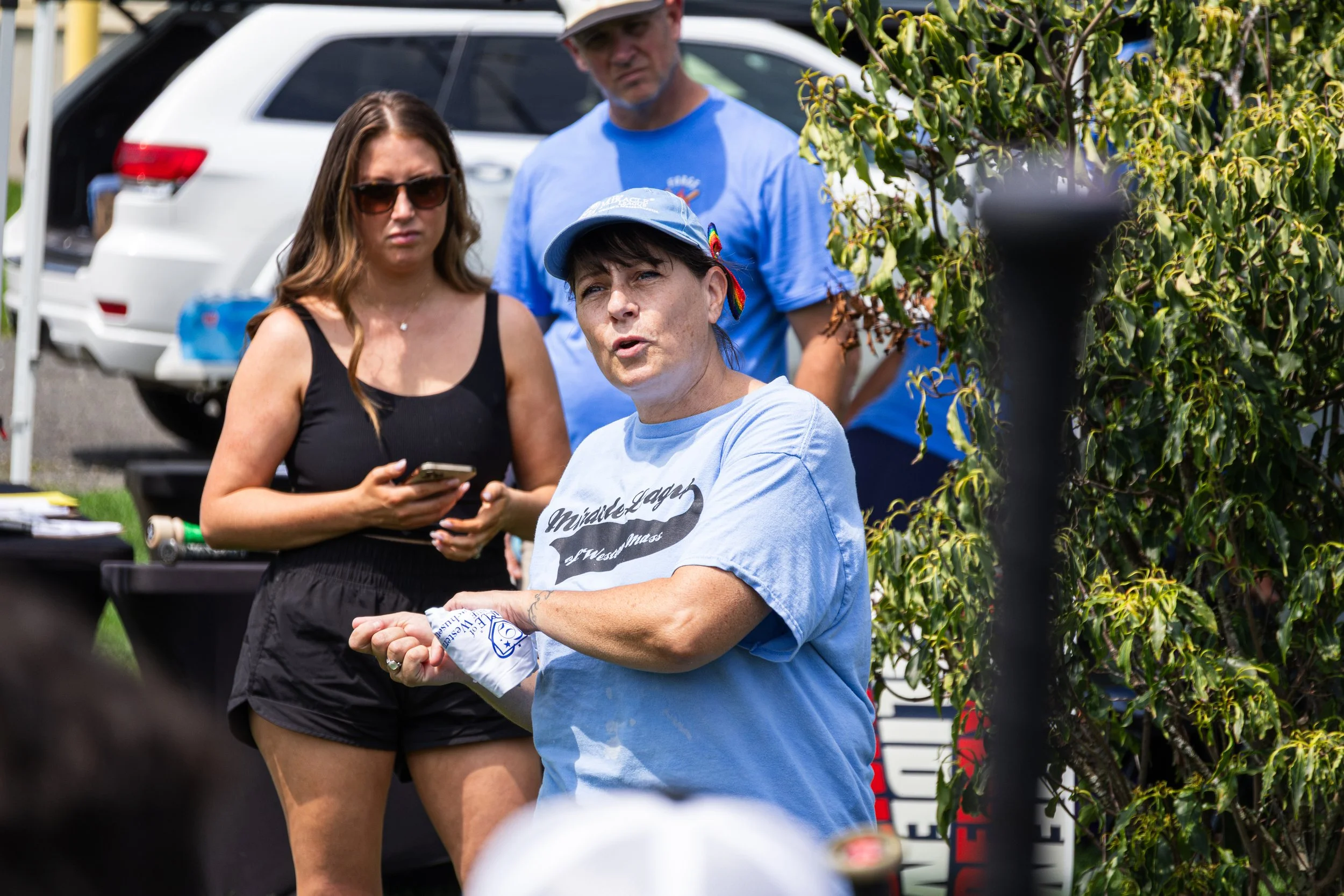Three women are engaged in a conversation outdoors near parked cars. One woman, wearing a black tank top and sunglasses, is holding a phone. Another woman, with dark hair tied back, wearing a light blue shirt and cap, is speaking and gesturing with h