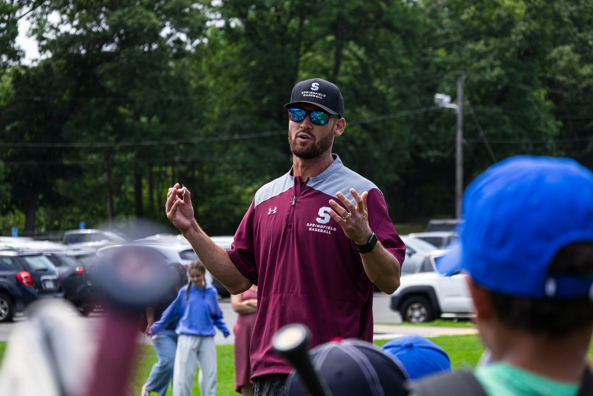 A man wearing a maroon Springfield Baseball shirt, black cap with a white 'S' and 'Springfield Baseball' on it, and reflective sunglasses, is speaking or instructing a group of children during an outdoor event with cars parked in the background and g