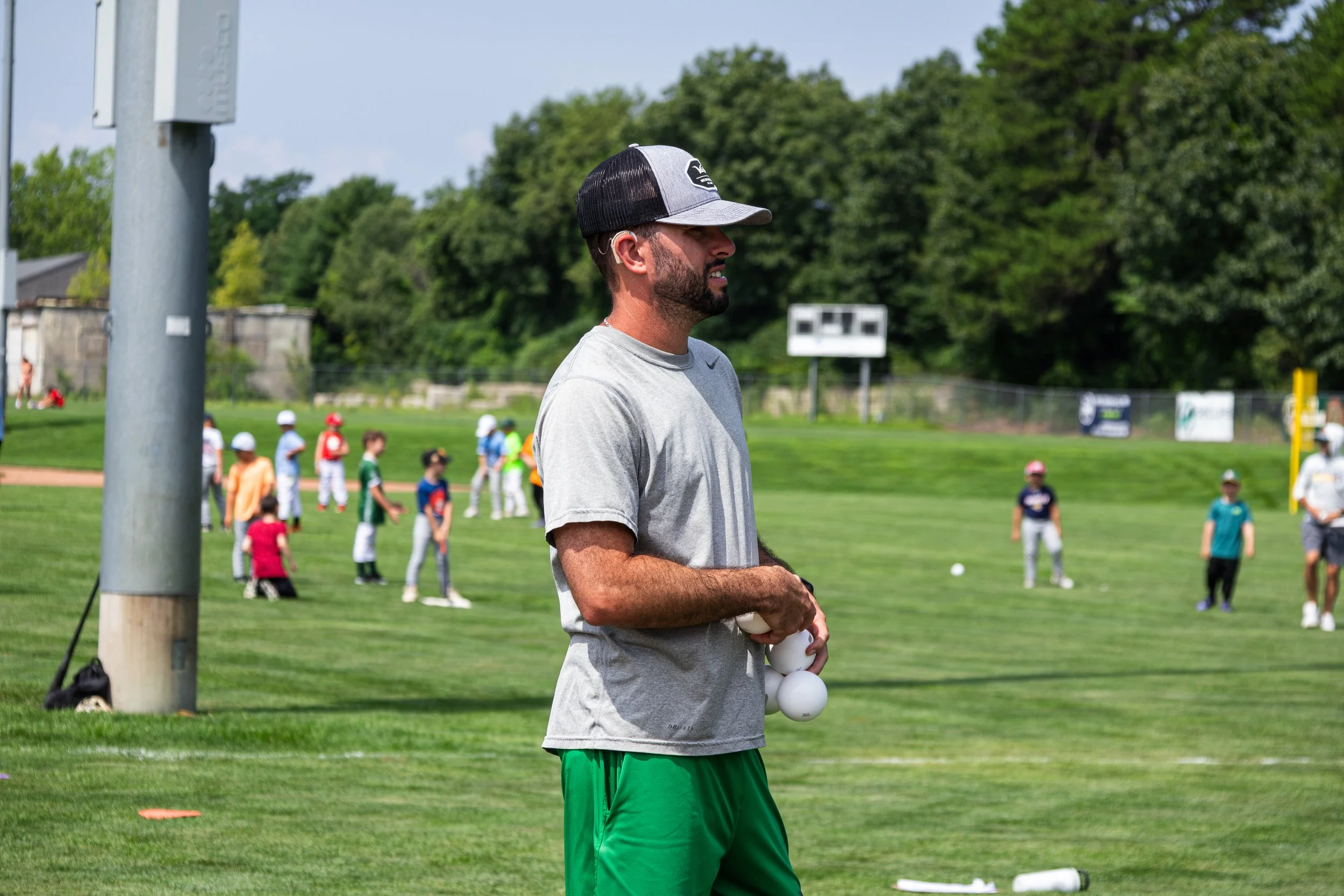 Man standing on a baseball field with children in the background, holding multiple white baseballs, wearing a gray T-shirt, green shorts, and a black and white cap.