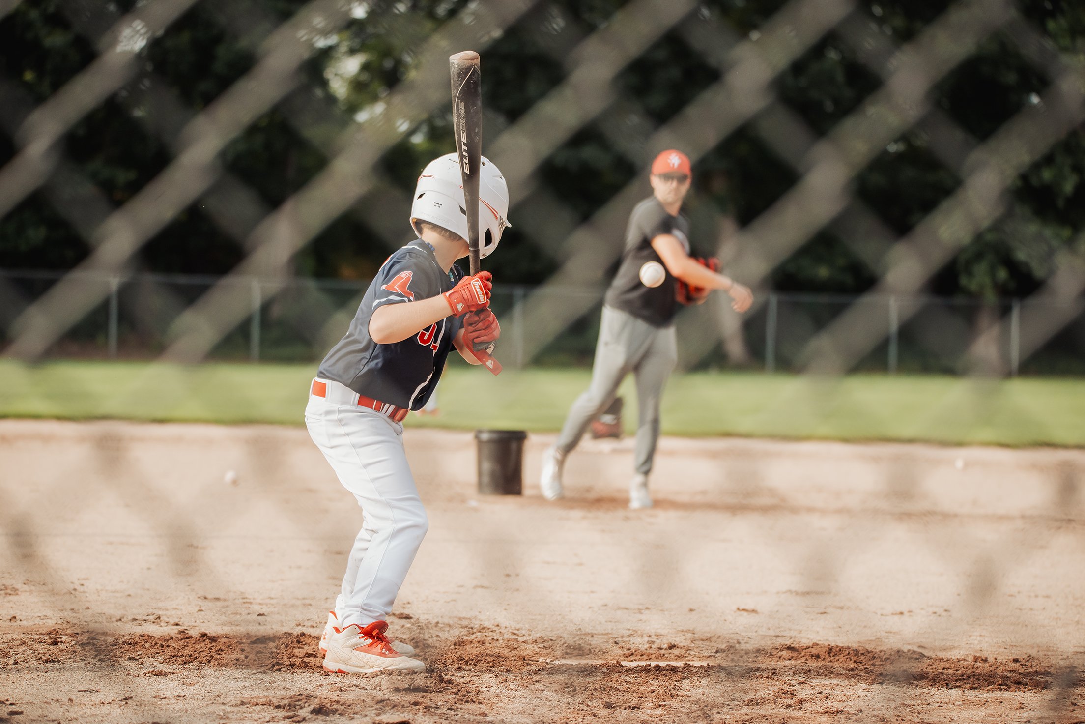 Young baseball player in batting stance at home plate, wearing helmet and glove, while teammate in background prepares to catch a baseball on a baseball field.