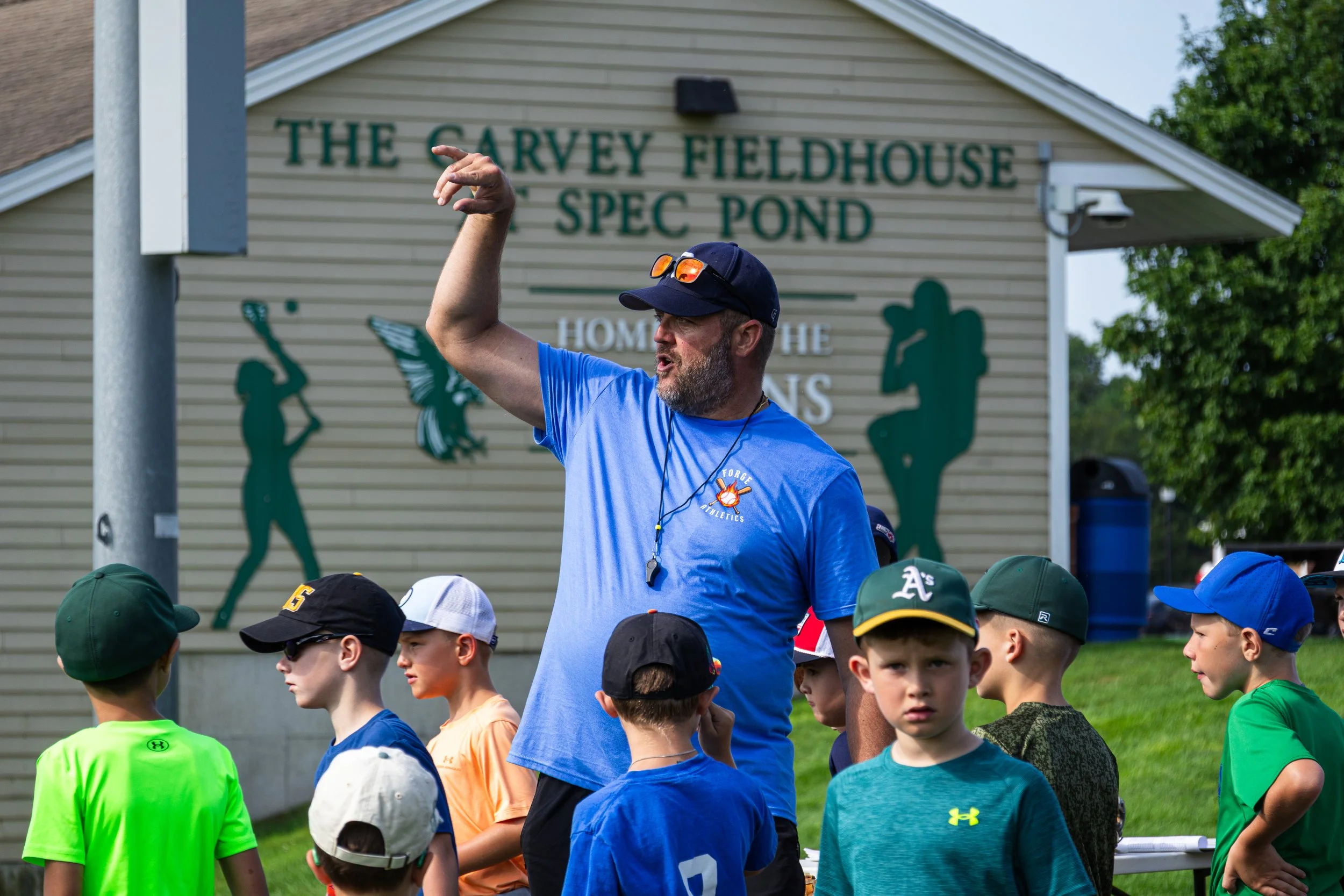 A man wearing a blue shirt and a dark cap is speaking to a group of children outdoors. The children are wearing colorful sports shirts and caps. In the background, there is a building with a sign that reads 'The Carvey Fieldhouse & Spec Pond' and som