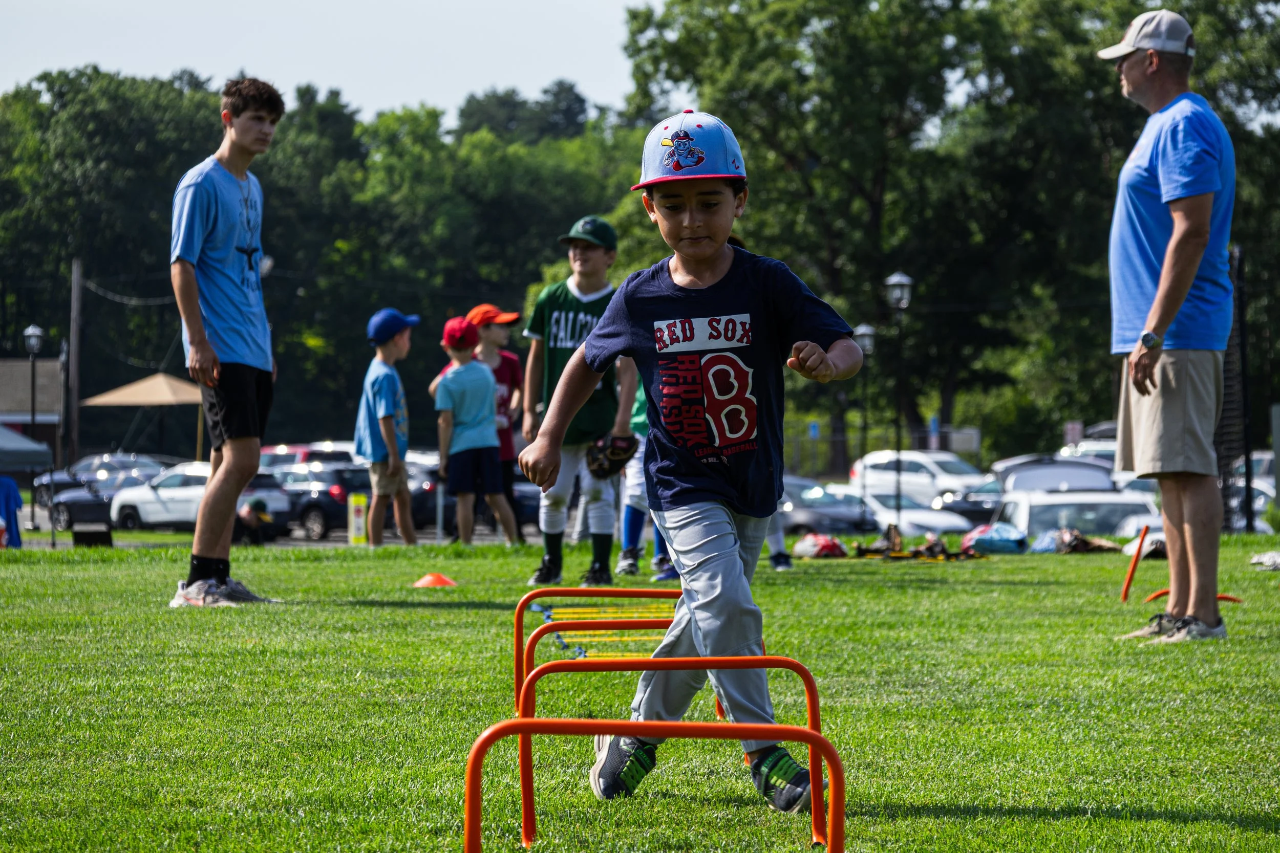 Young boy wearing a blue baseball cap and dark T-shirt participating in an outdoor athletic activity, jumping over orange hurdles, with other children and adults in the background during a sunny day at a park or sports field.
