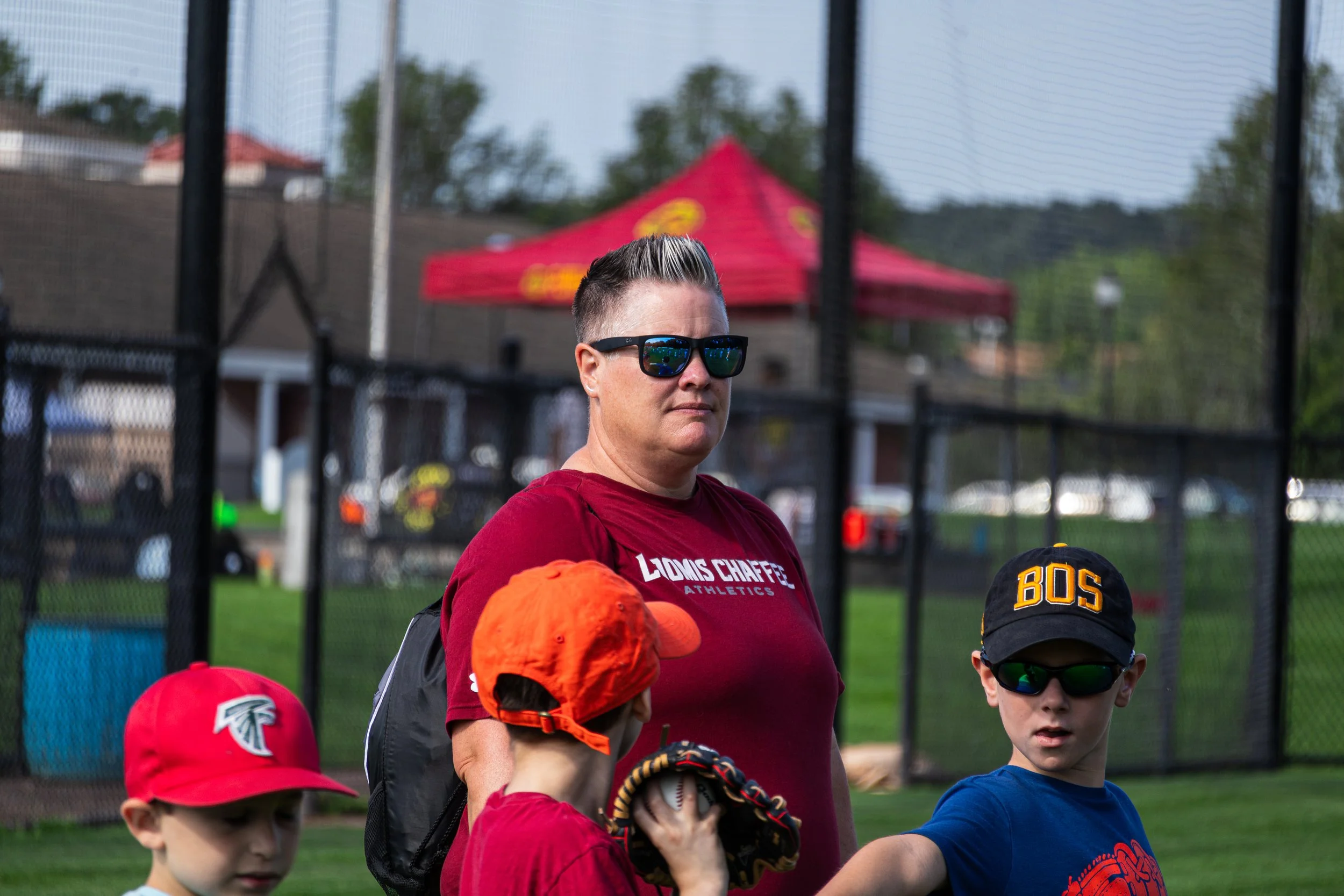 A woman wearing sunglasses and a red shirt stands behind a group of children, some wearing Atlanta Falcons hats, as they prepare for a baseball game.